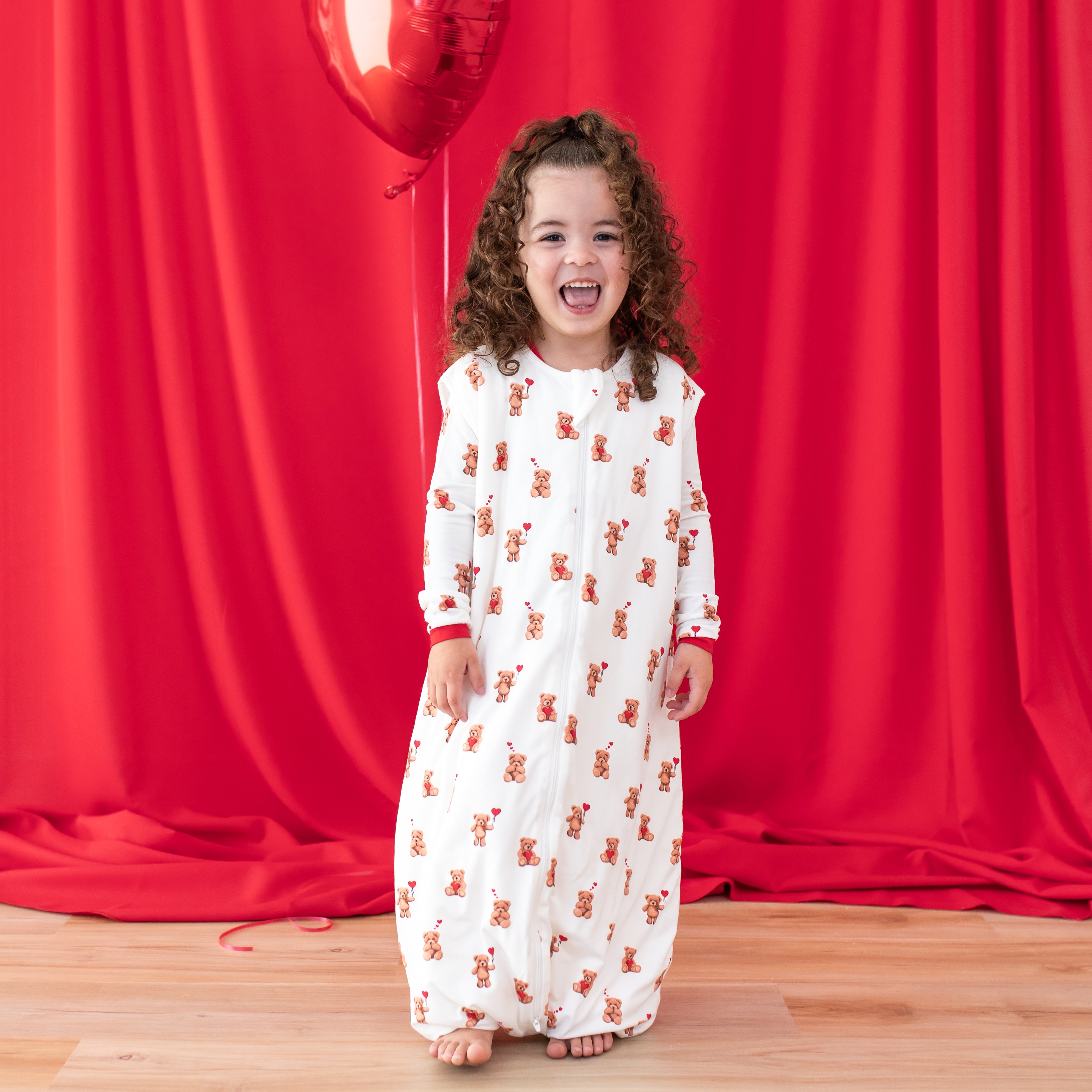 Smiling young girl standing in front of a red curtain wearing the soft and breathable Sleep Bag Walker in Bear Hearts 1.0 with matching long sleeve pajamas on underneath