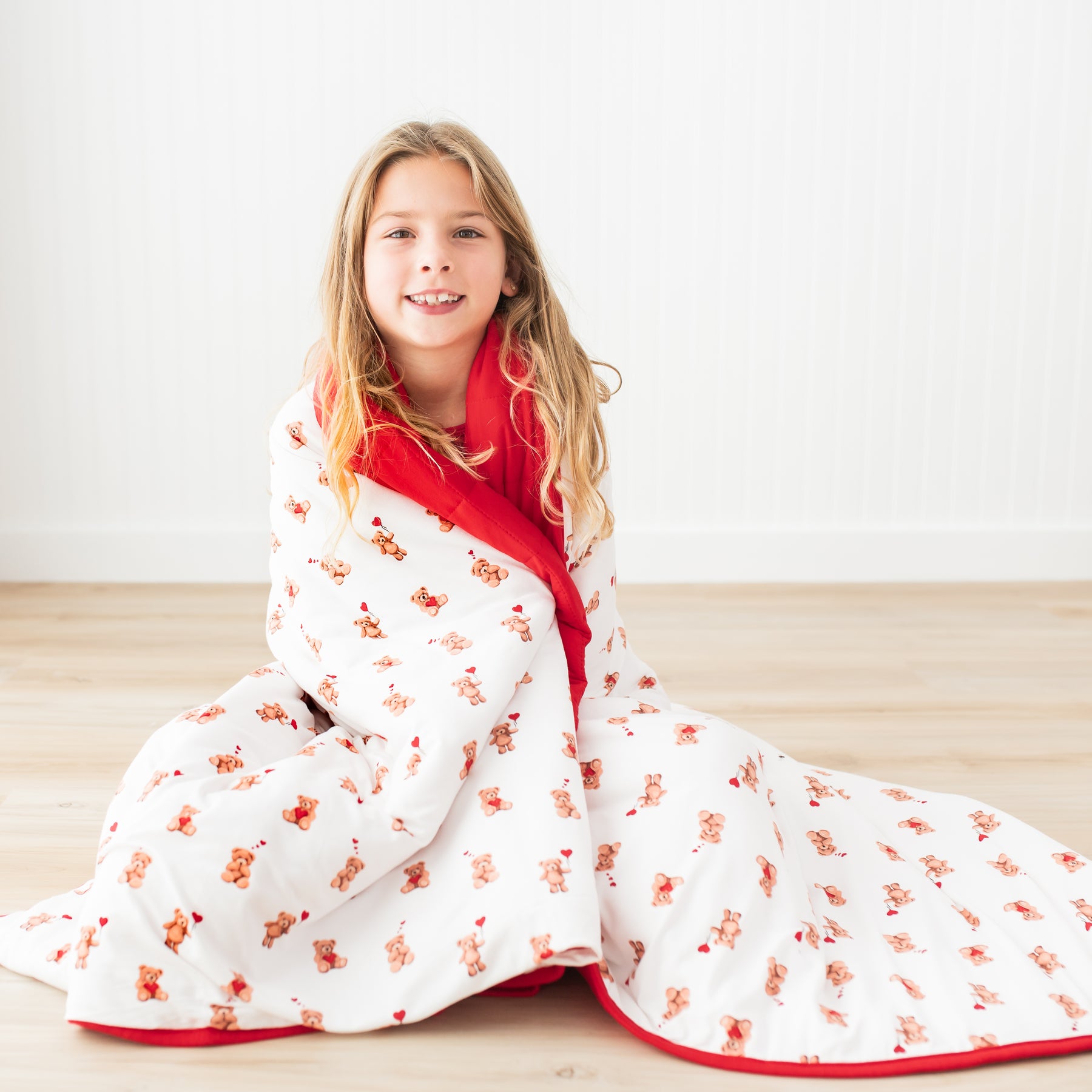 Young girl sitting on the wooden floor in front of a white paneled wall with the Youth Blanket in Bear Hearts 2.5 wrapped around her shoulders