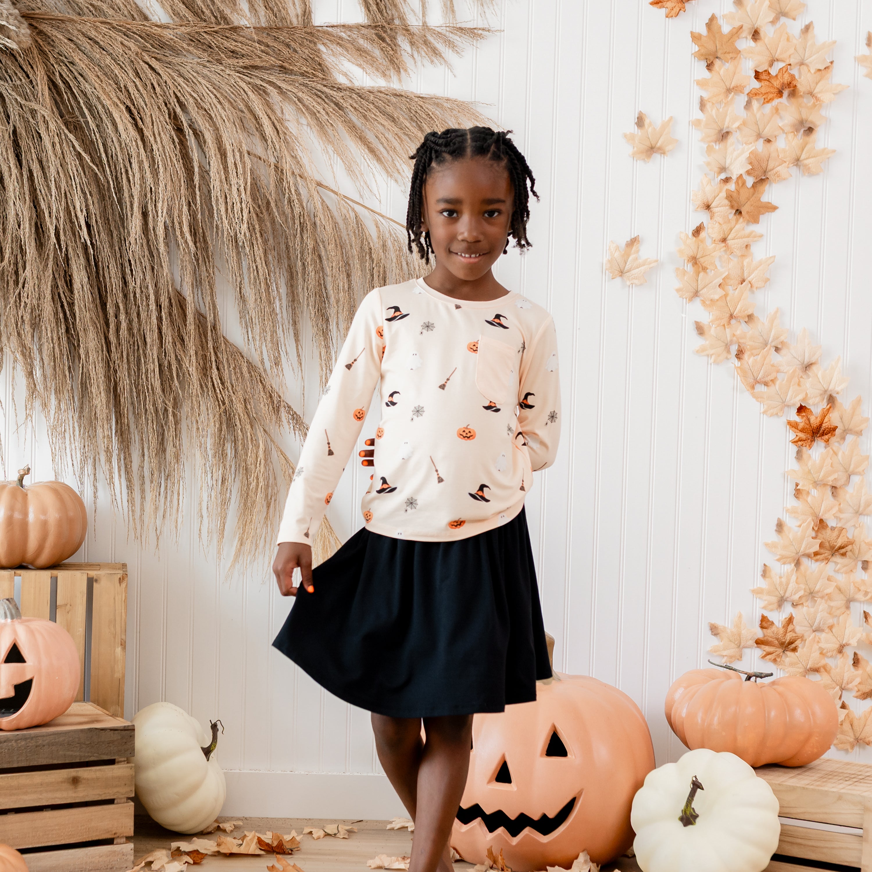 Young girl standing wearing the Long Sleeve Toddler Crew Neck Tee in Bewitched and Midnight skort in front of a fall themed backdrop