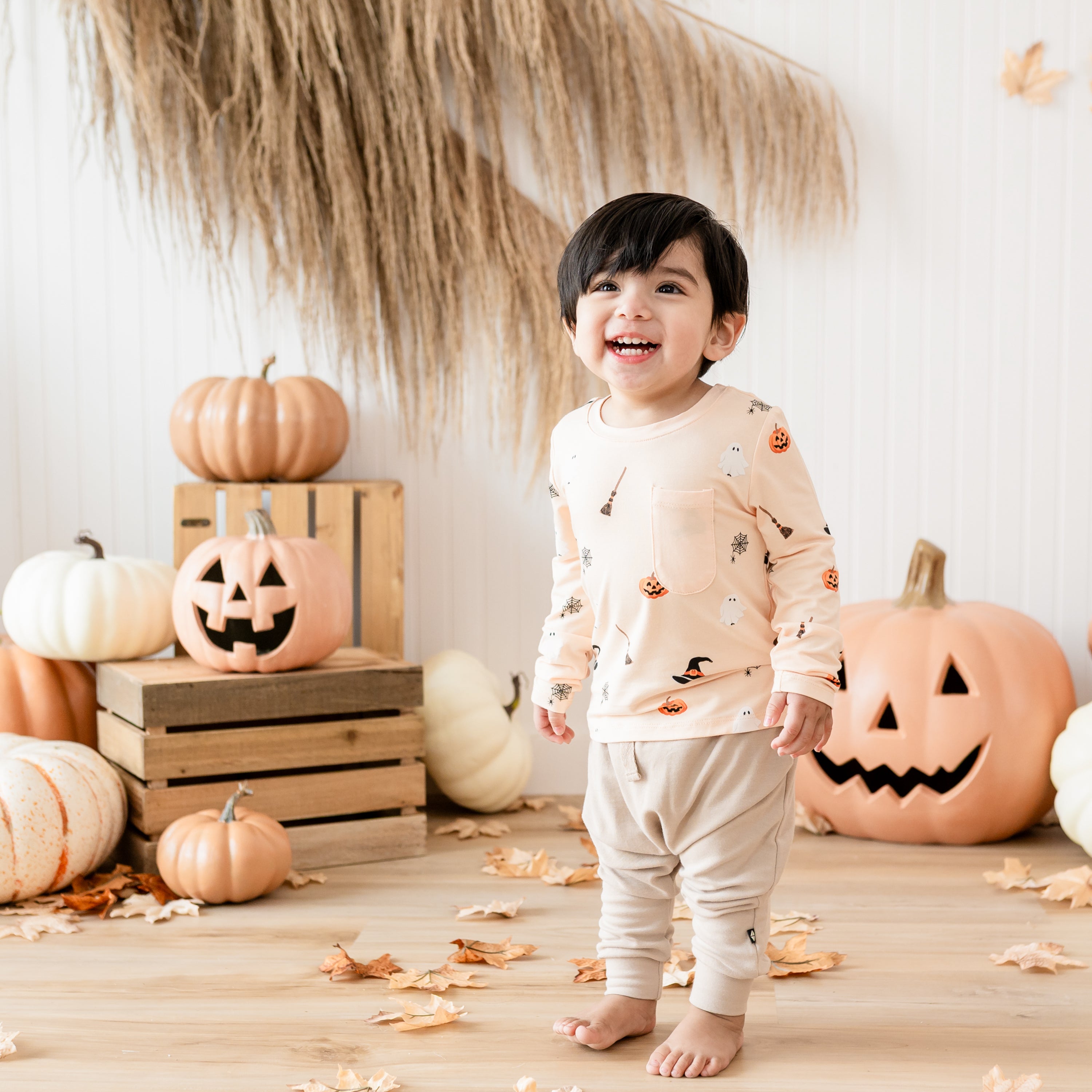 Young toddler smiling up at the camera wearing the Long Sleeve Toddler Crew Neck Tee in Bewitched paired with Bamboo Jersey Harem pants in front of a rustic fall inspired, Halloween backdrop