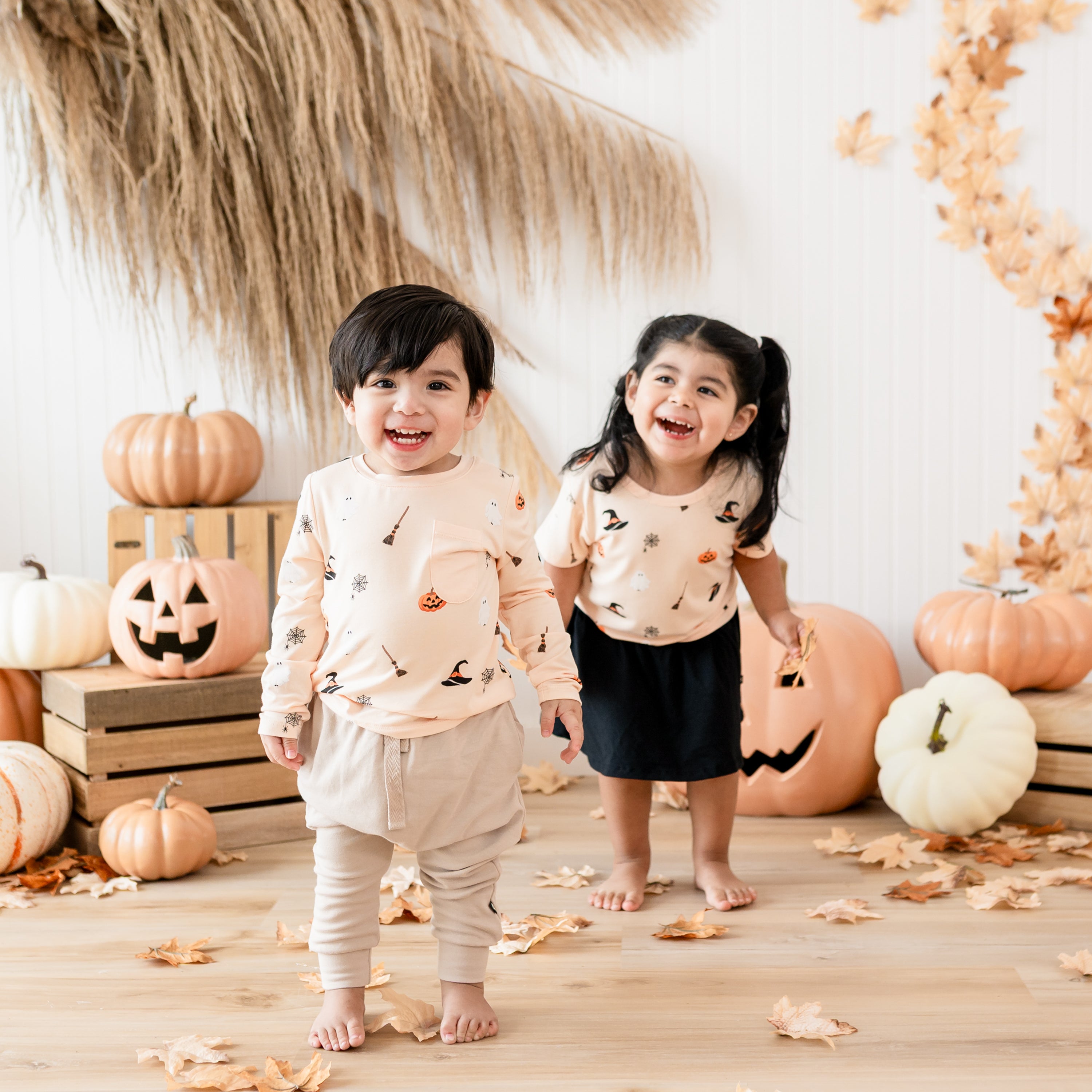 Two young toddler wearing the Long Sleeve Toddler Crew Neck Tee and basic tee in Bewitched standing in front of a rustic fall inspired Halloween backdrop 