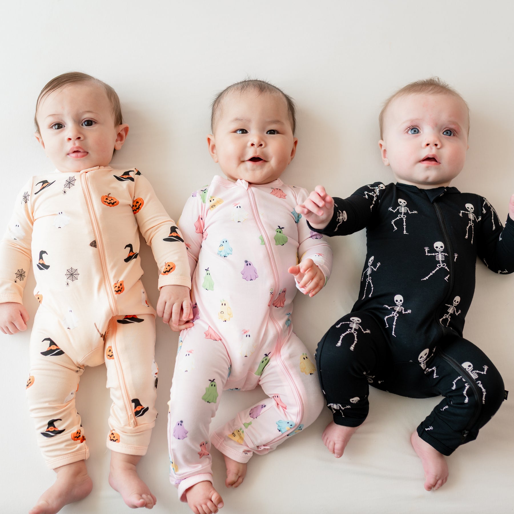 Three infants laying side by side on a light neutral background wearing the Zippered Romper in Bewitched, Pastel Ghosts and Skeleton