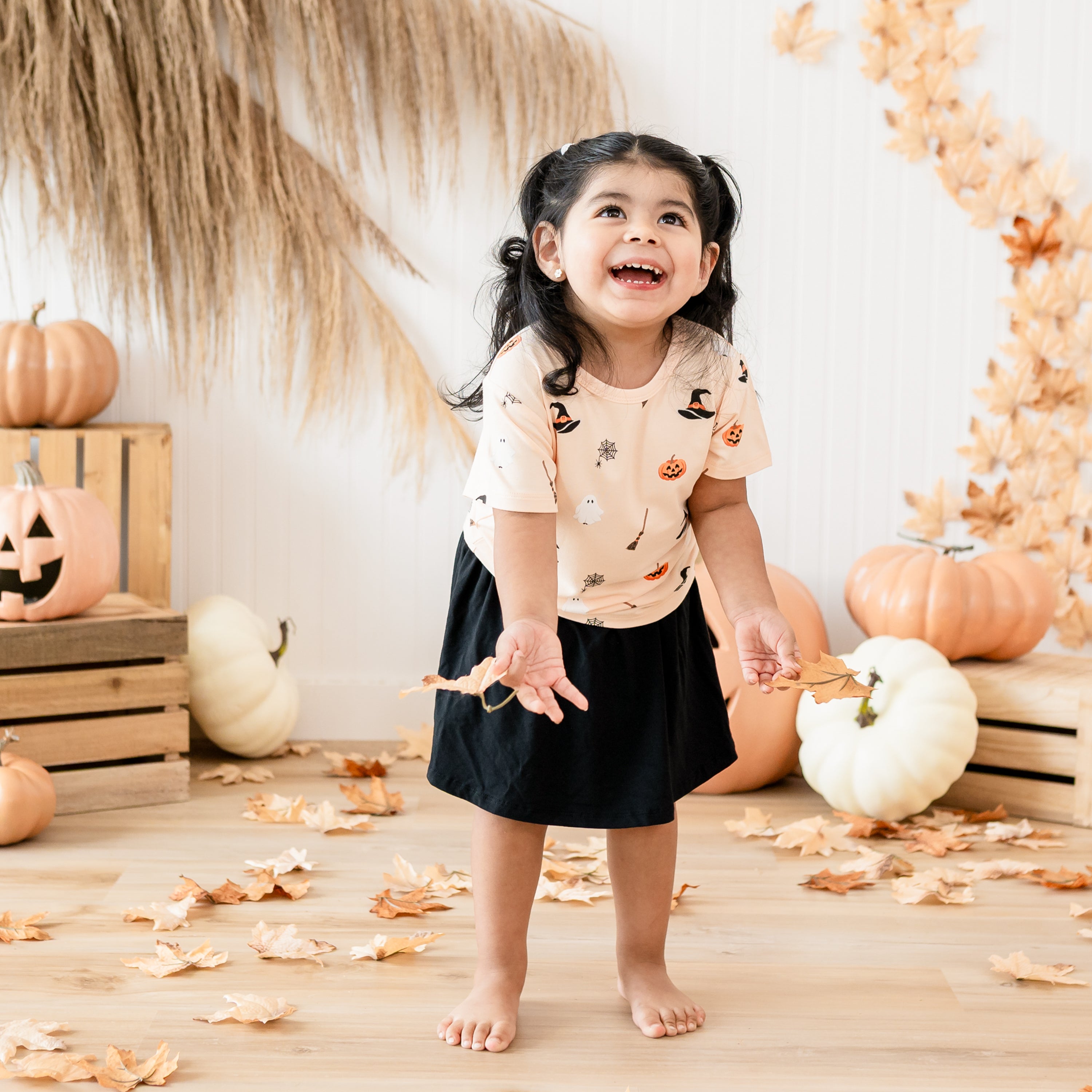 Smiling toddler looking up wearing the Toddler Basic Tee in Bewitched paired with a midnight skort holding leaves in both her hands in front of a fall inspired back drop