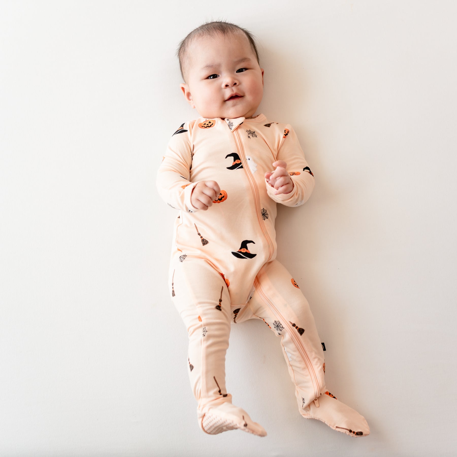 Infant laying on a light neutral background wearing the Zippered Footie in Bewitched