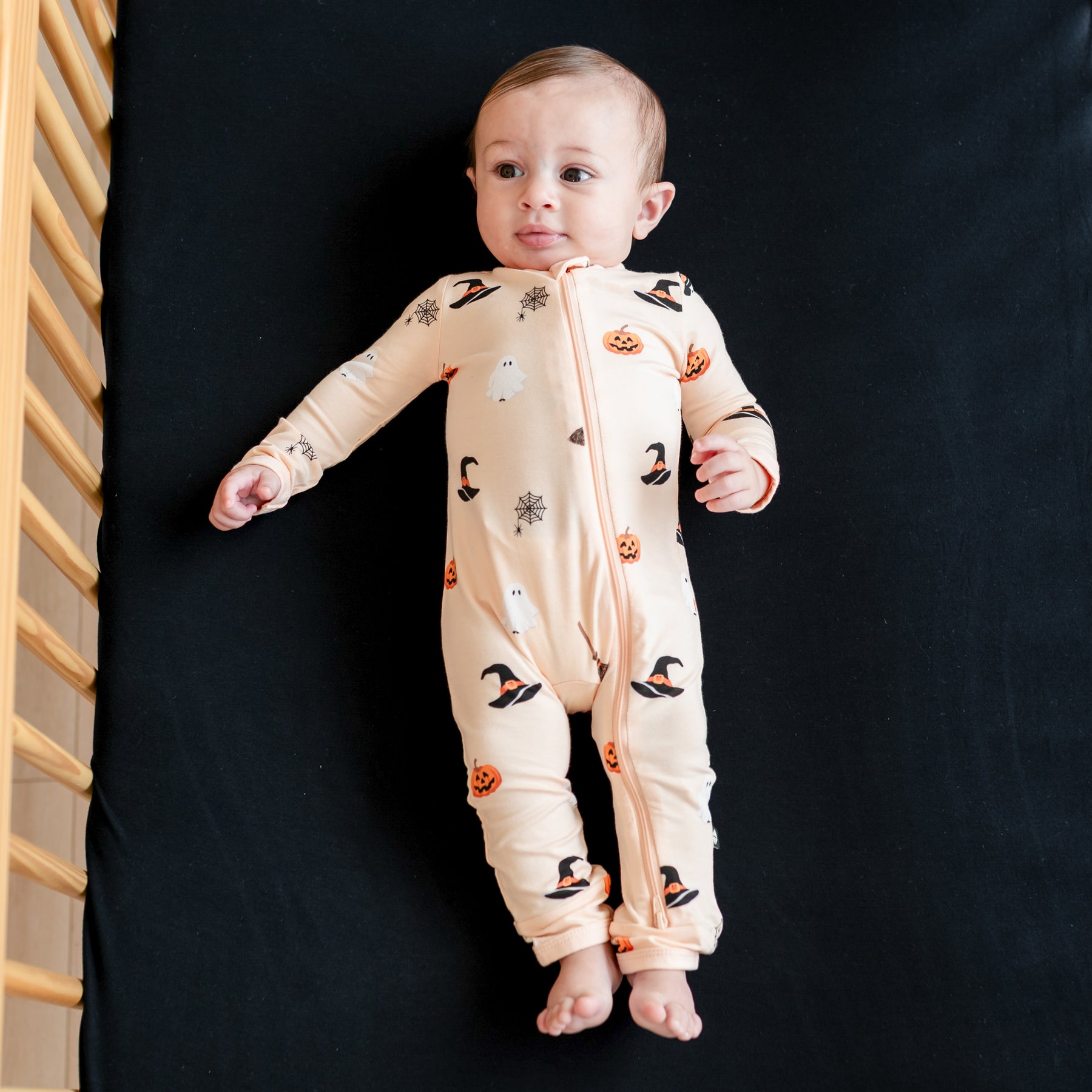 Infant laying in a crib on a midnight crib sheet wearing the Zippered Romper in Bewitched