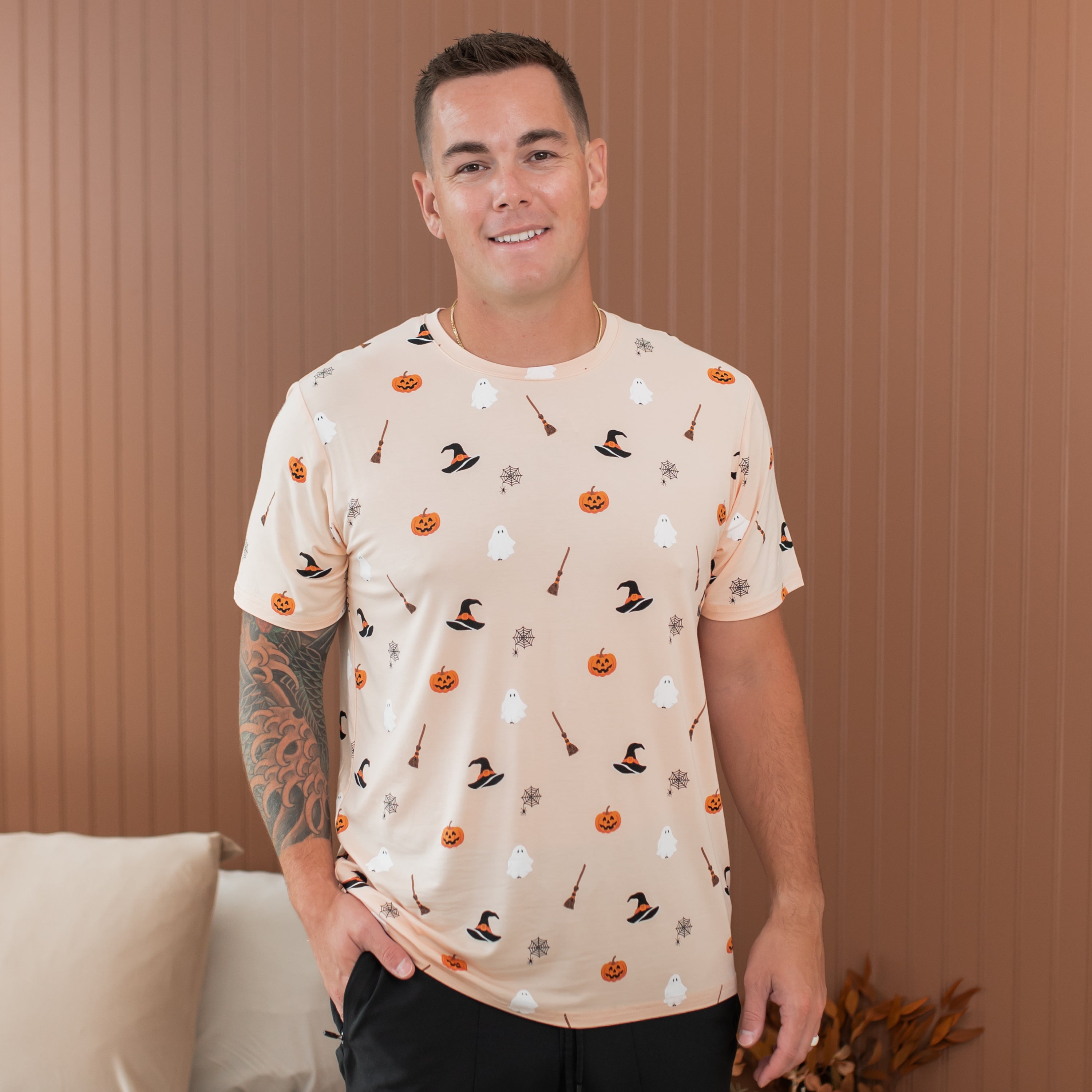 Man wearing a Halloween-themed t-shirt with pumpkins, witches' hats, and brooms against a brown background.