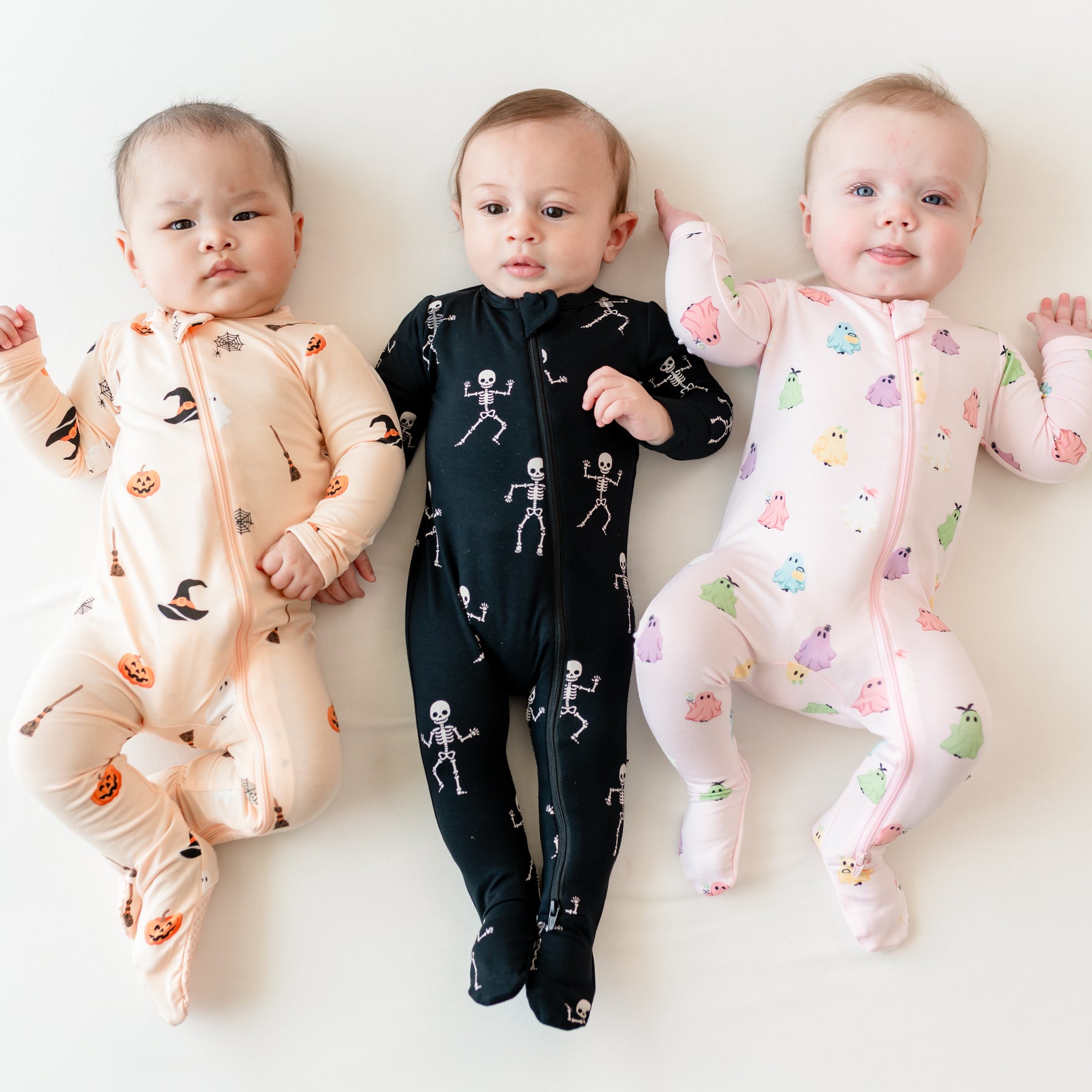 Three infant models laying side by side on a light neutral background wearing Zippered Footie in Bewitched, Skeleton and Pastel Ghosts