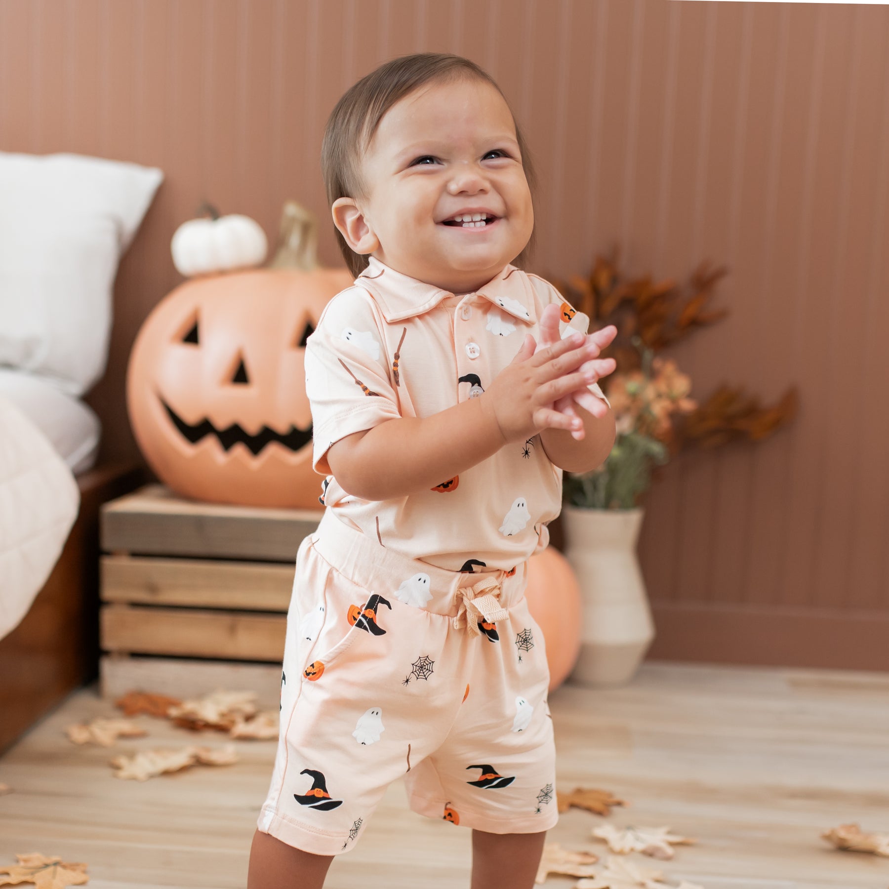 Child in a Halloween-themed matching polo shirt and shorts clapping in front of pumpkins and autumn decorations.