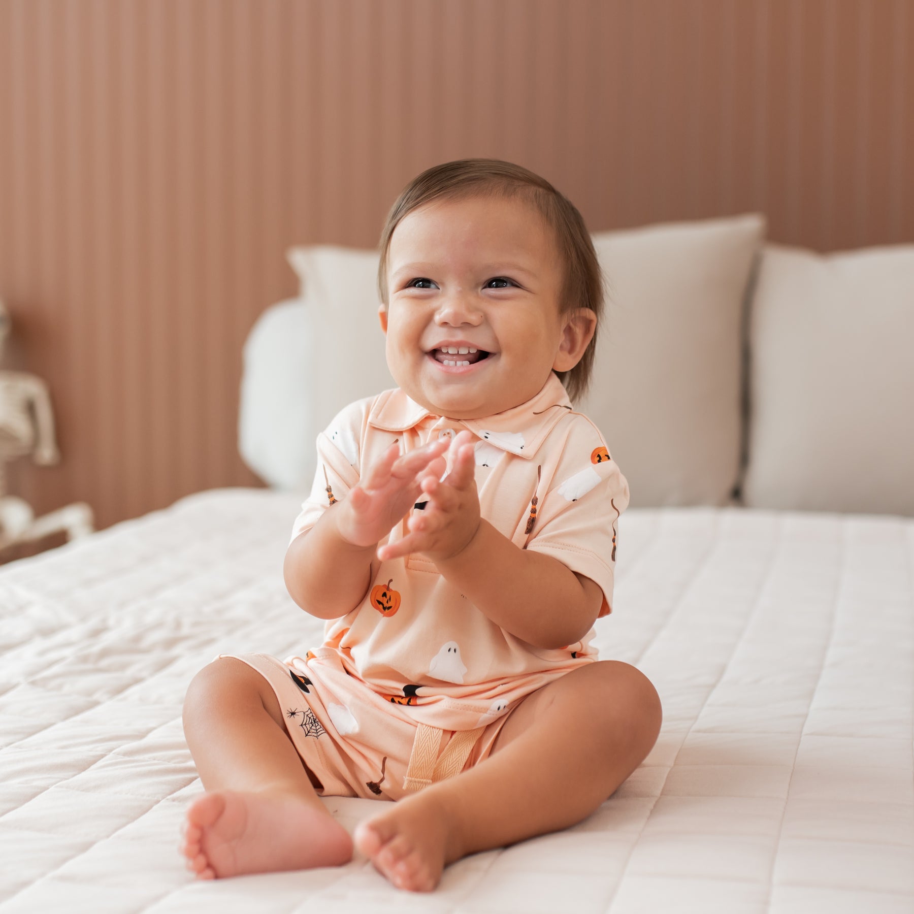 Baby in a halloween outfit sitting on a bed clapping hands with a smile 