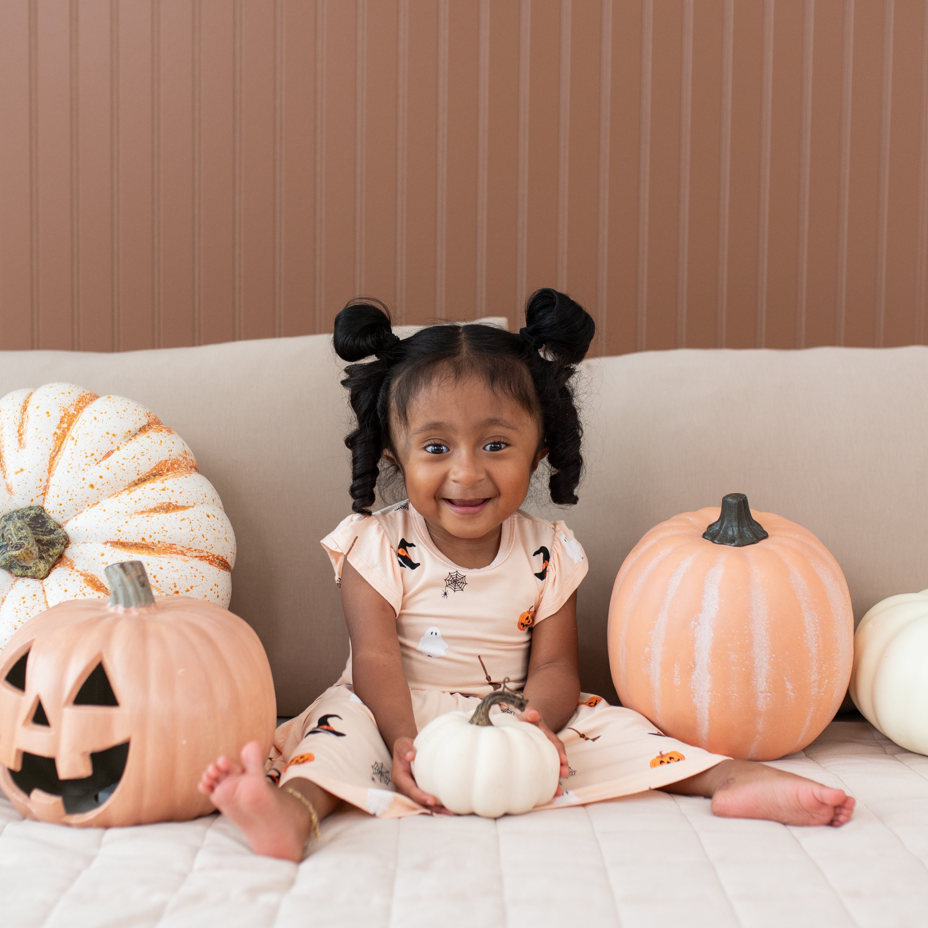 Child sitting on a couch with pumpkins, wearing a Halloween-themed dress.