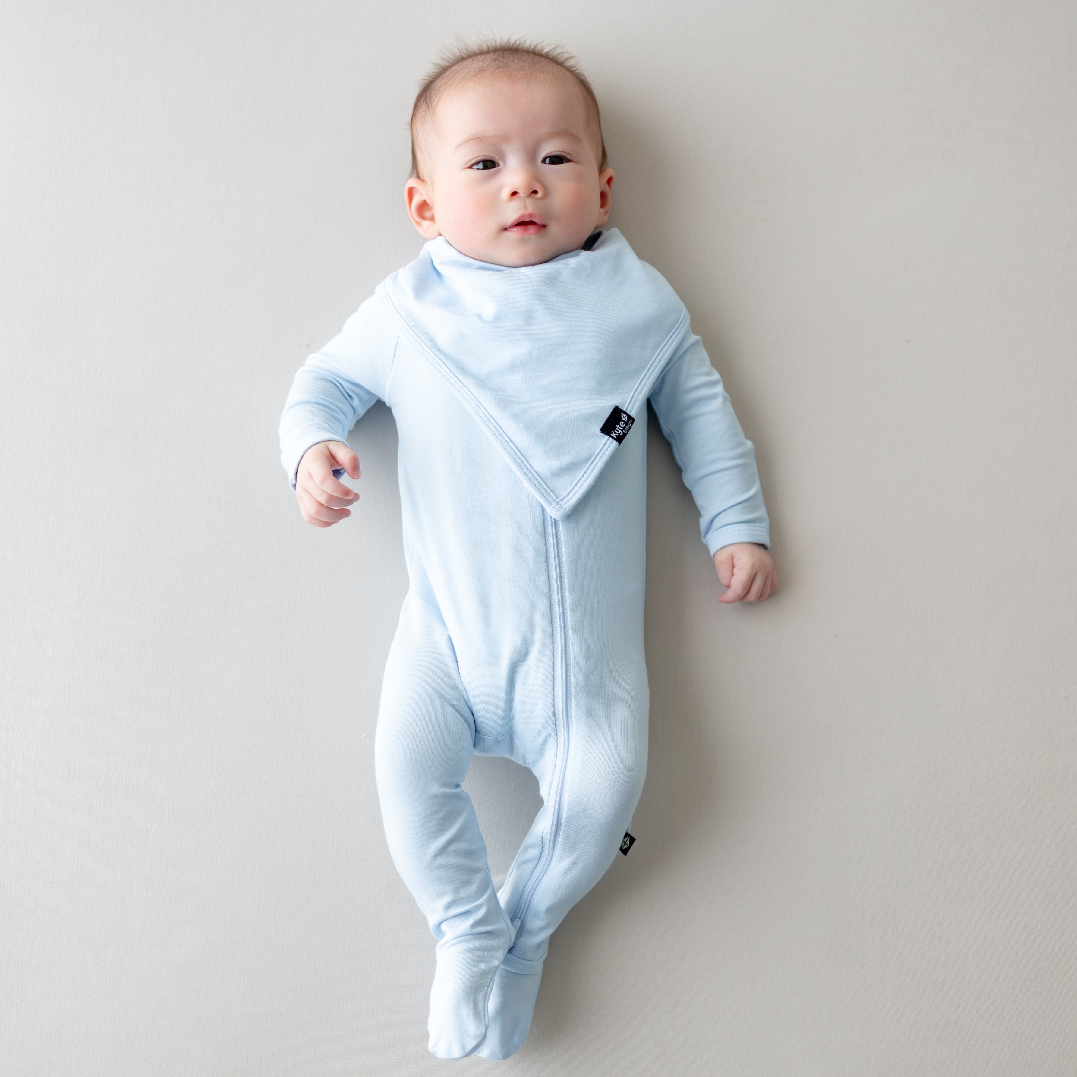 Infant laying on a light neutral surface wearing the Bib in Breeze overtop a matching zippered footie