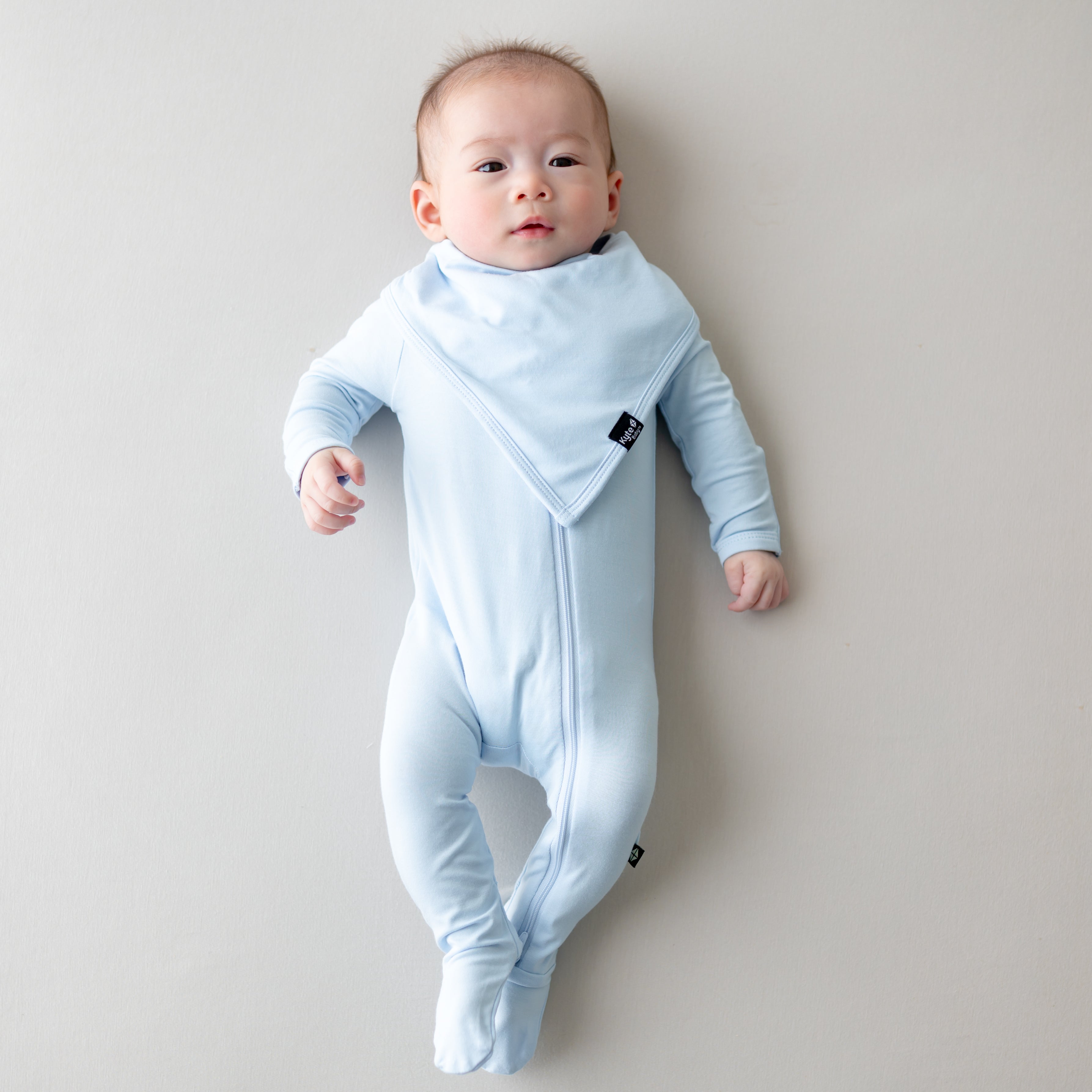 Infant laying on a light neutral surface wearing the Bib in Breeze overtop a matching zippered footie