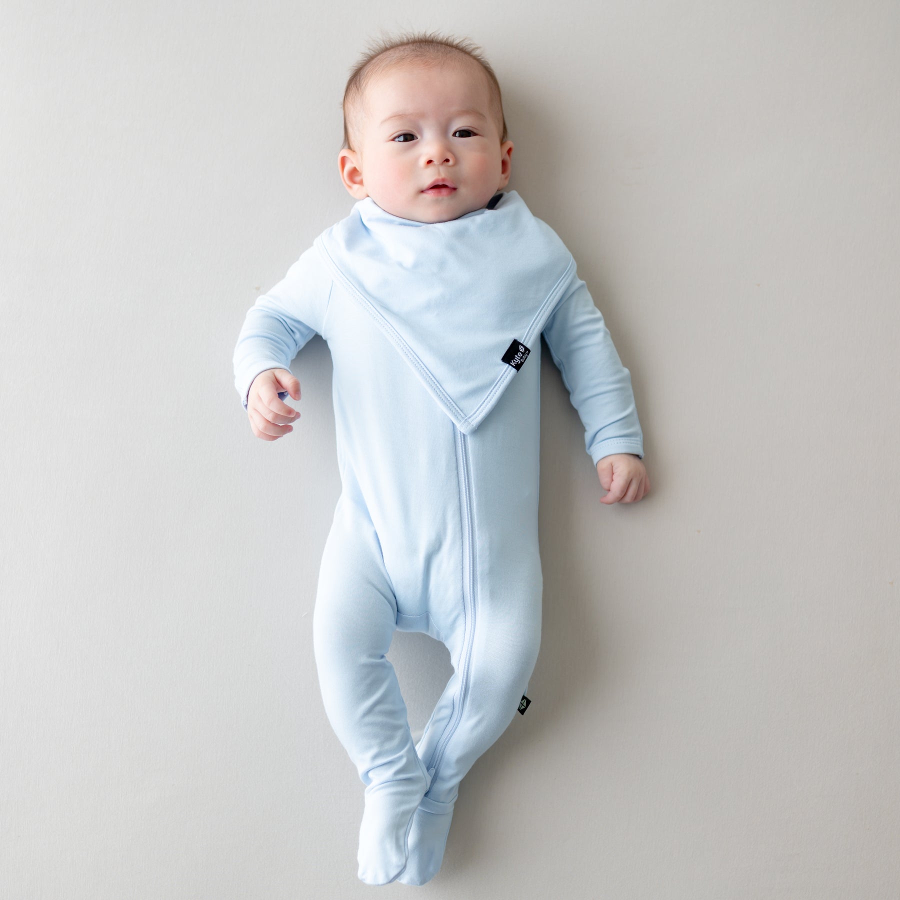 Infant laying on a light neutral surface wearing the Bib in Breeze overtop a matching zippered footie