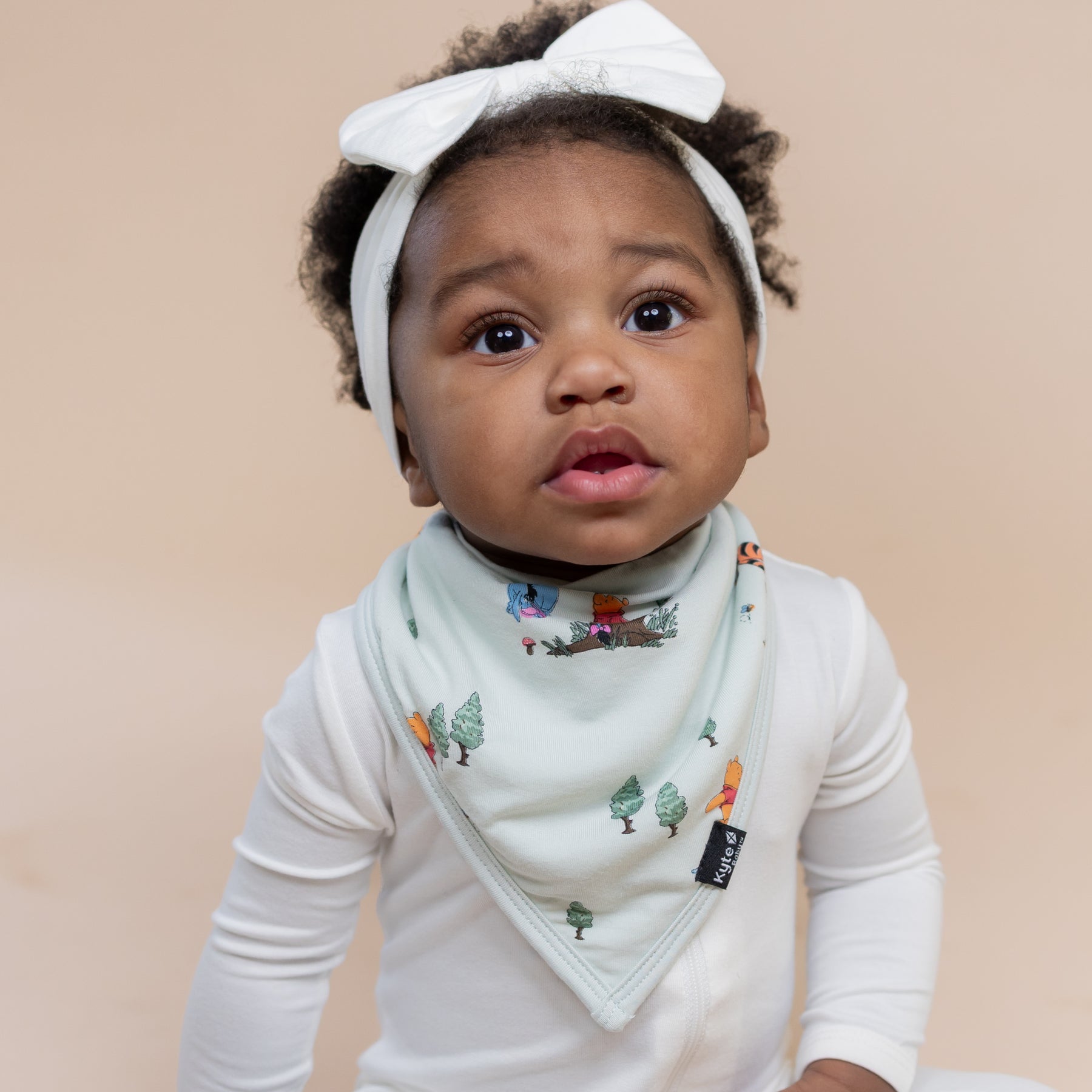 Young toddler wearing the Bib in Winnie the Pooh Hundred Acre Wood over a white zippered romper paired with a white bow