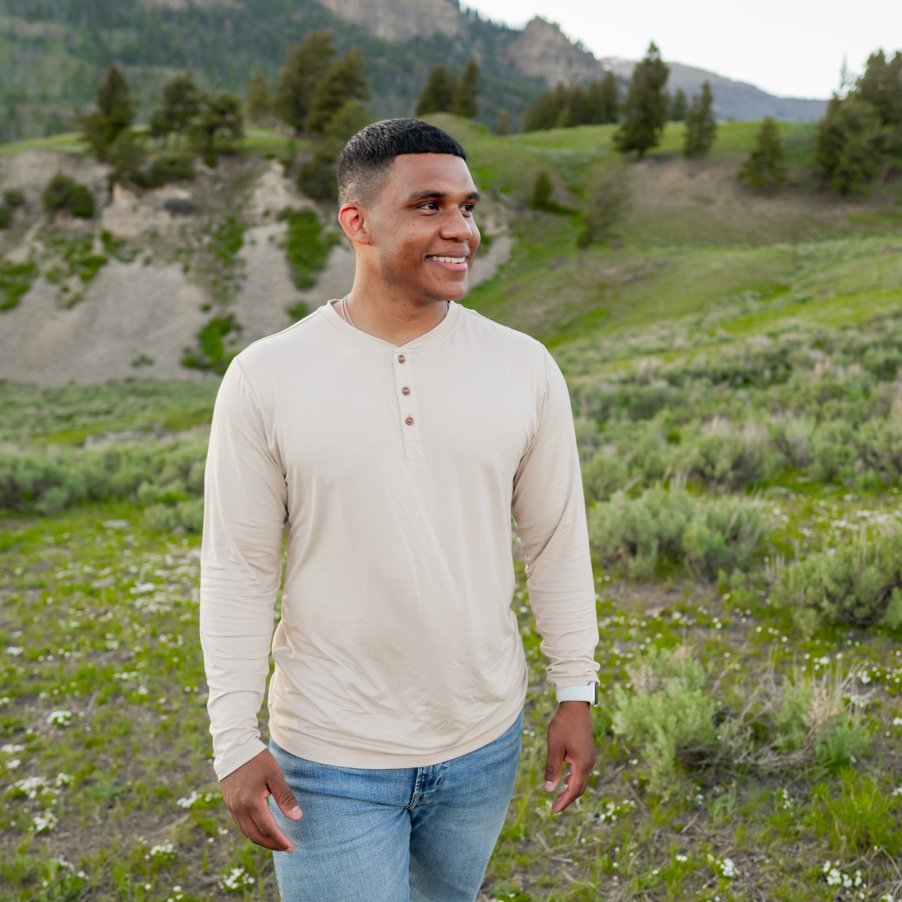 Smiling male model standing in a field outside wearing the Men's Long Sleeve Henley Top in Bisque