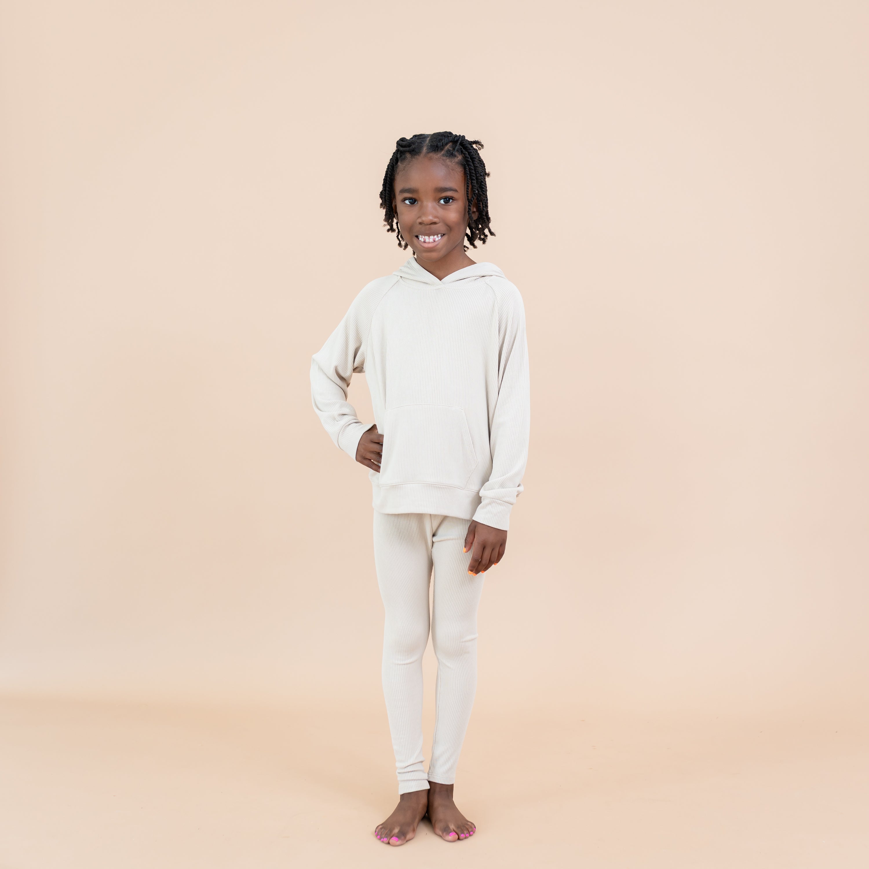 Smiling girl standing with one hand on her hip wearing the Ribbed Leggings in Bisque paired with a matching ribbed hoodie in front of a light taupe background