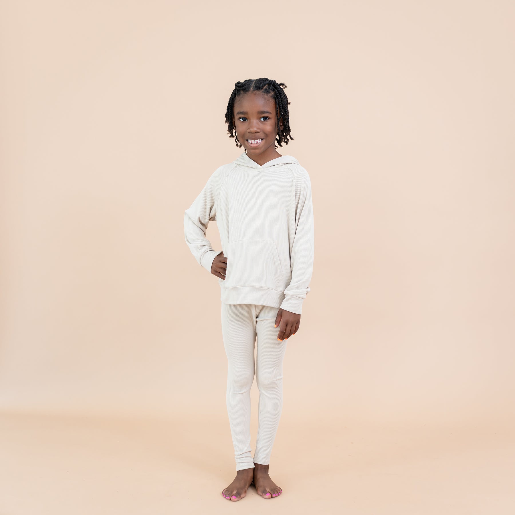 Smiling girl standing with one hand on her hip wearing the Ribbed Leggings in Bisque paired with a matching ribbed hoodie in front of a light taupe background