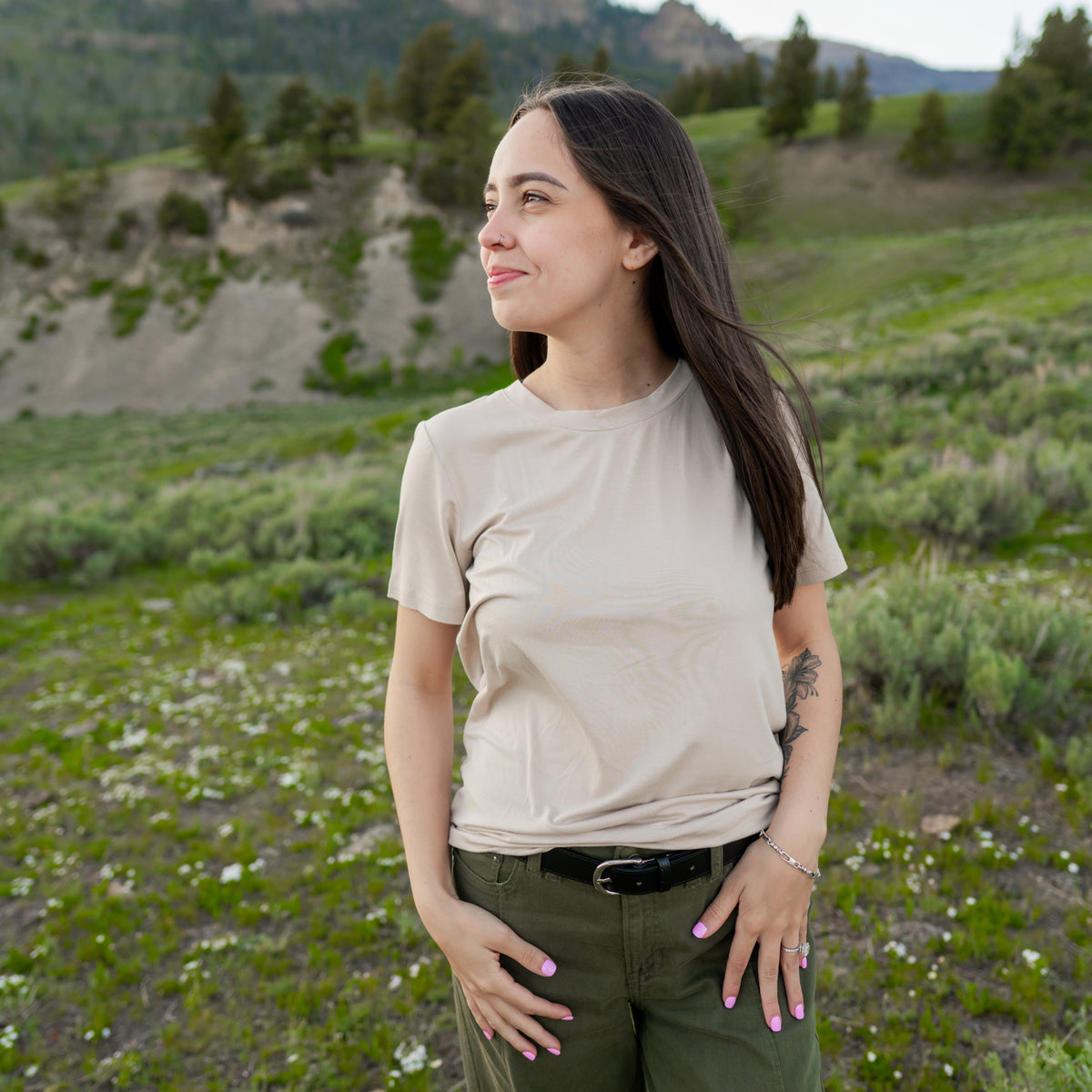 Smiling model standing in a field wearing the Women's Crew Neck Tee in Bisque paired with olive green pants