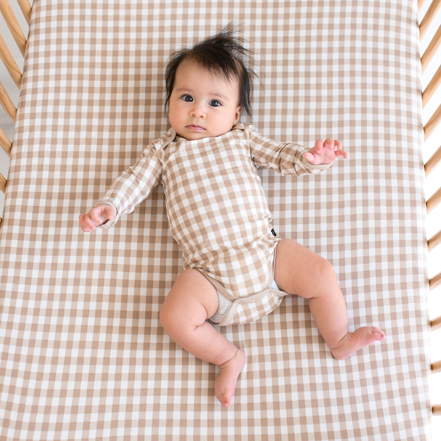 Infant girl laying in a crib wearing the Long Sleeve Bodysuit in Gingham Bisque on a matching crib sheet