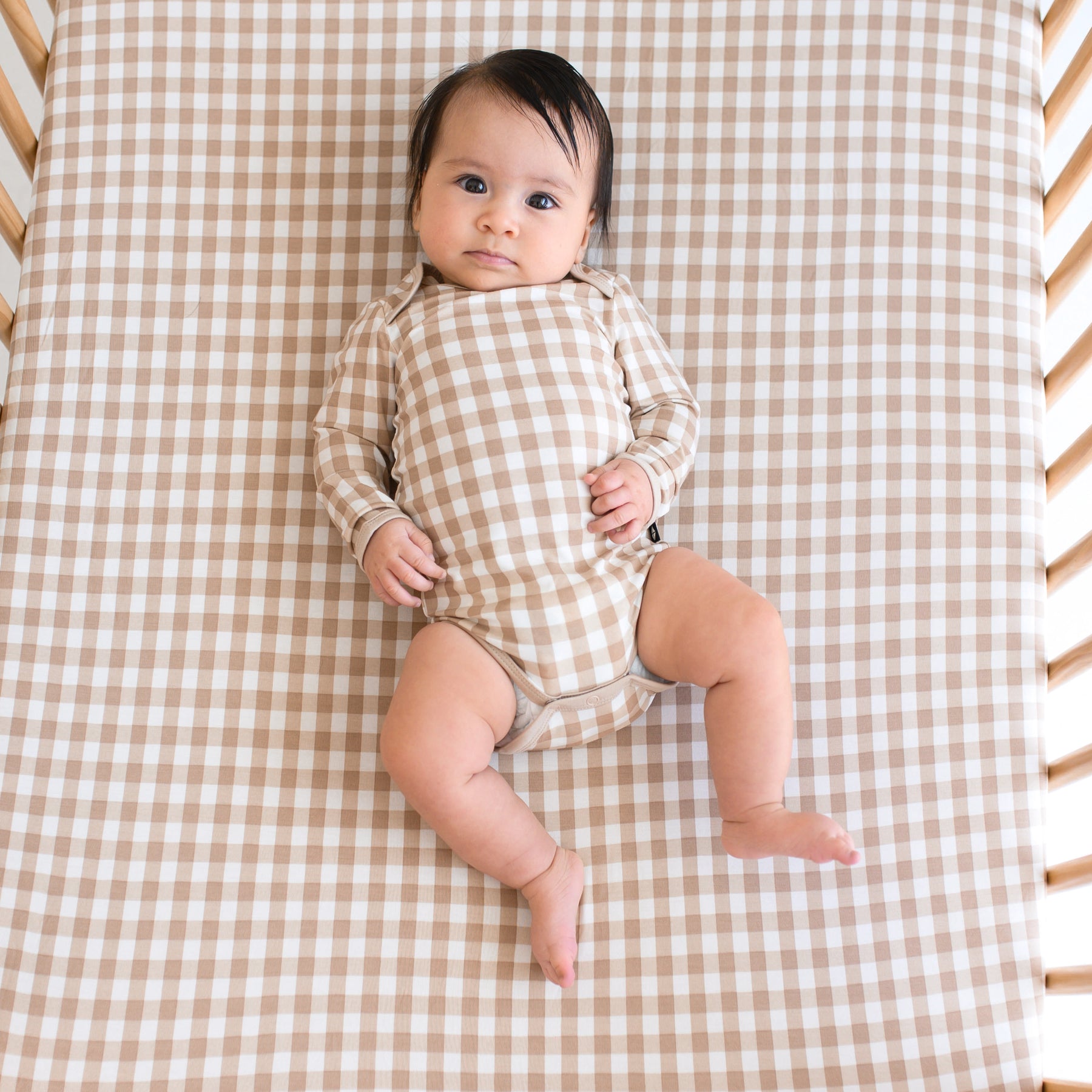 Infant girl laying in a crib wearing the Long Sleeve Bodysuit in Gingham Bisque on a matching crib sheet