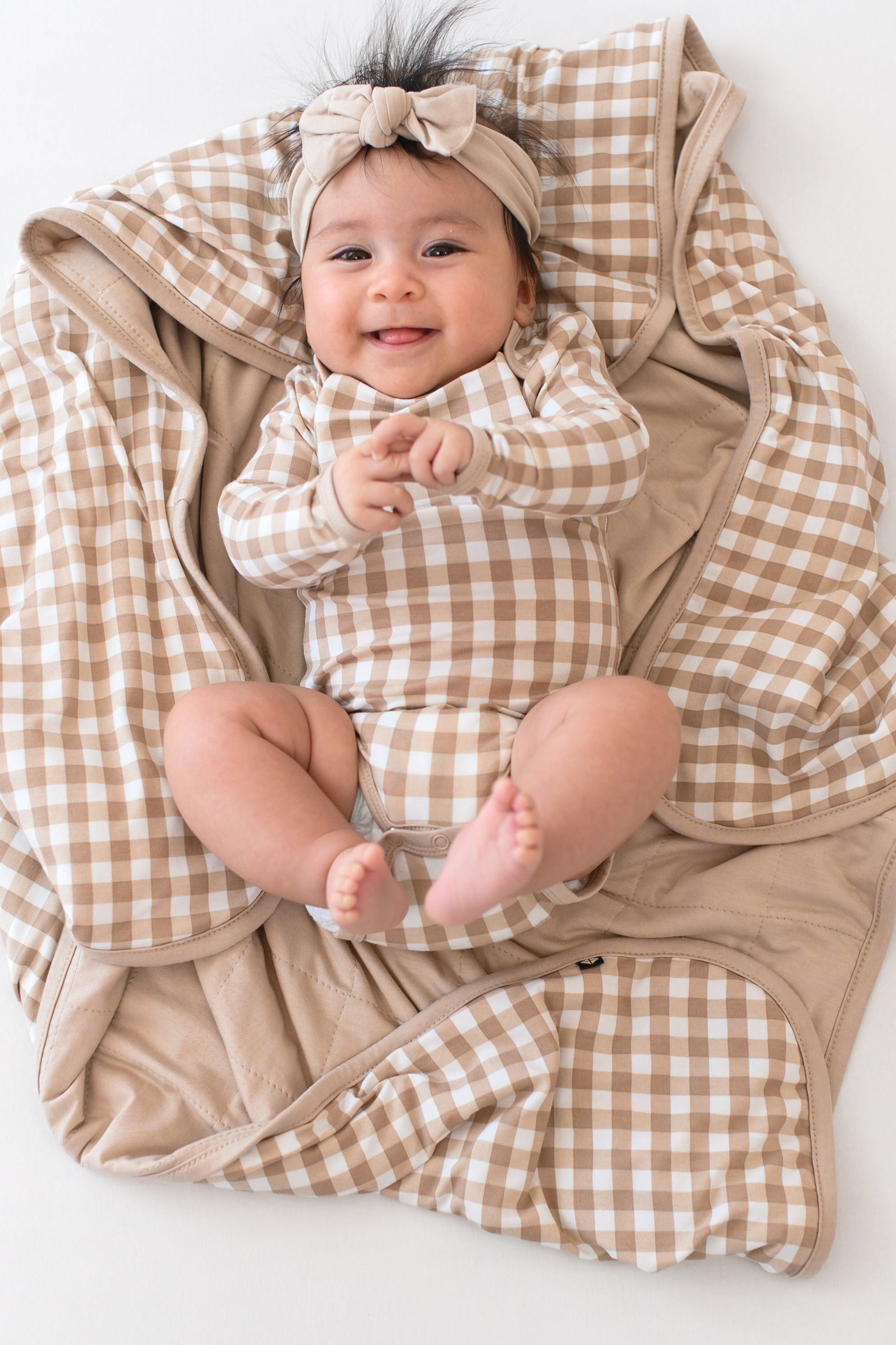 Infant laying on a Baby Blanket in Gingham Bisque wearing a matching long sleeve bodysuit and bisque knotted bow headband