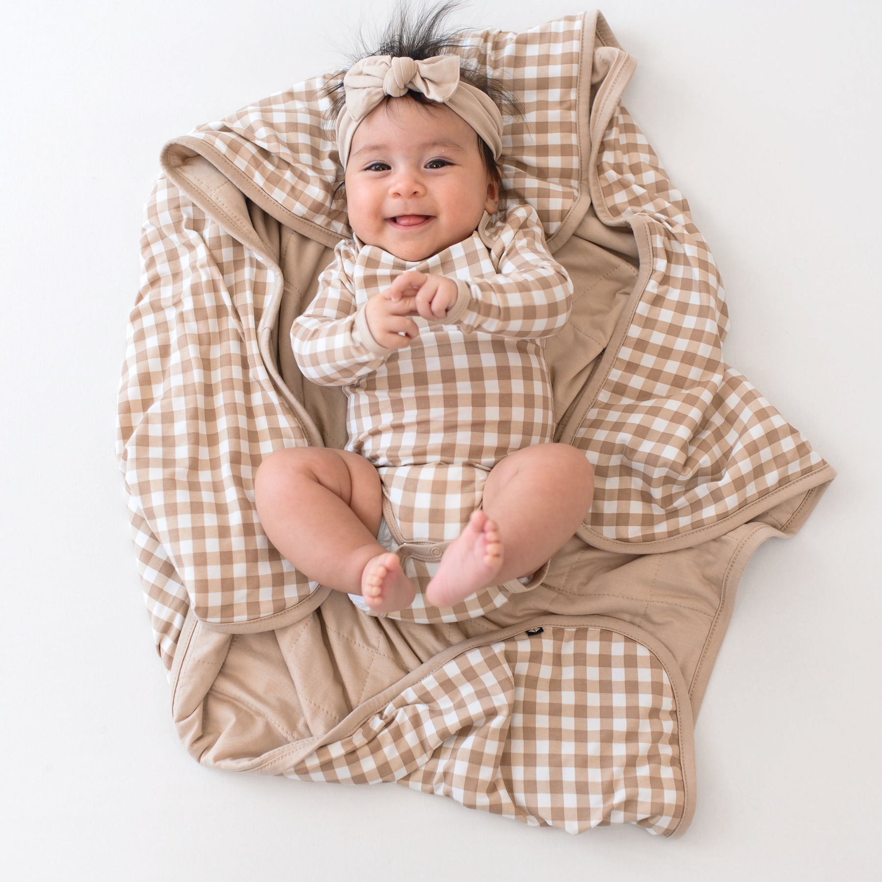 Infant laying on a Baby Blanket in Gingham Bisque wearing a matching long sleeve bodysuit and bisque knotted bow headband