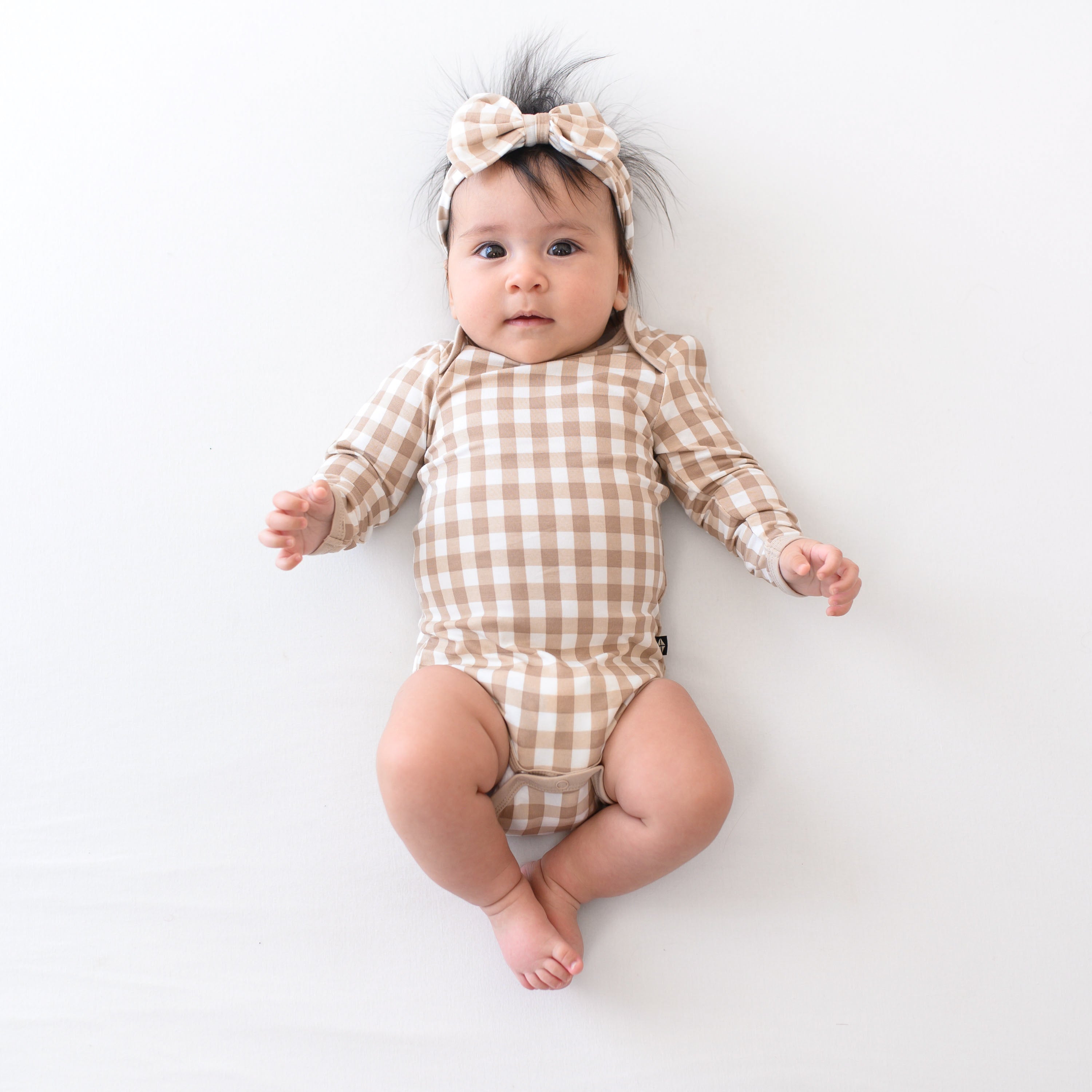 Infant girl wearing the Long Sleeve Bodysuit in Gingham Bisque with matching bow headband