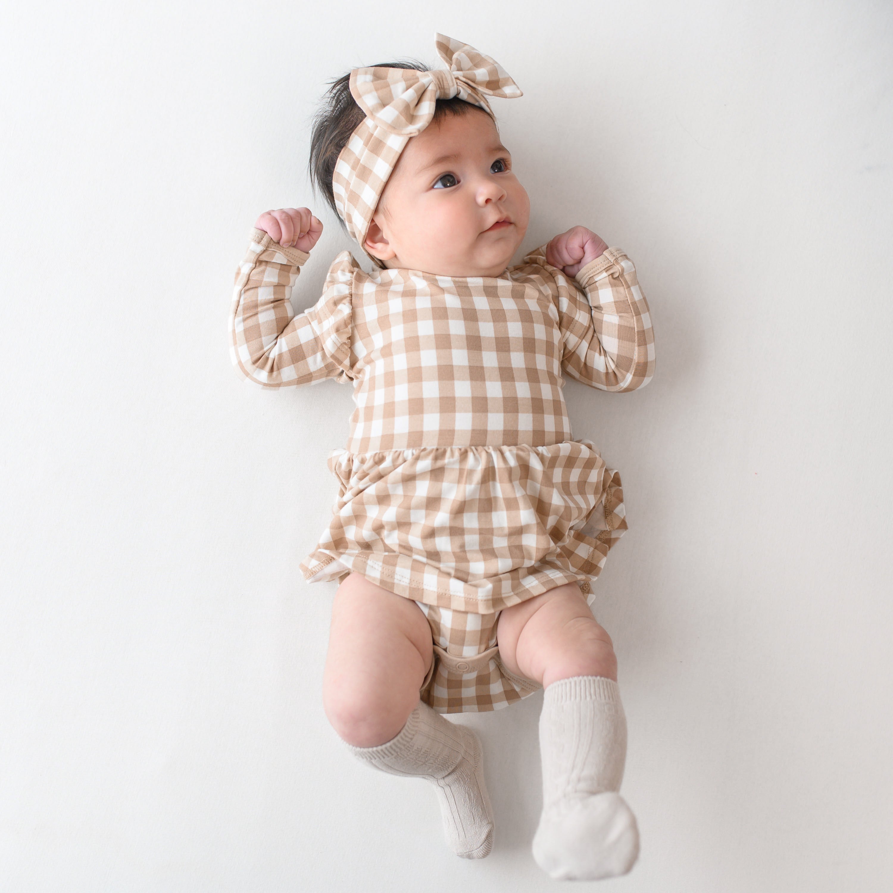 Infant girl laying on a light colored neutral backdrop looking off to the side wearing the Long Sleeve Twirl Bodysuit Dress in Gingham Bisque and matching bow headband with Oat Knee high socks