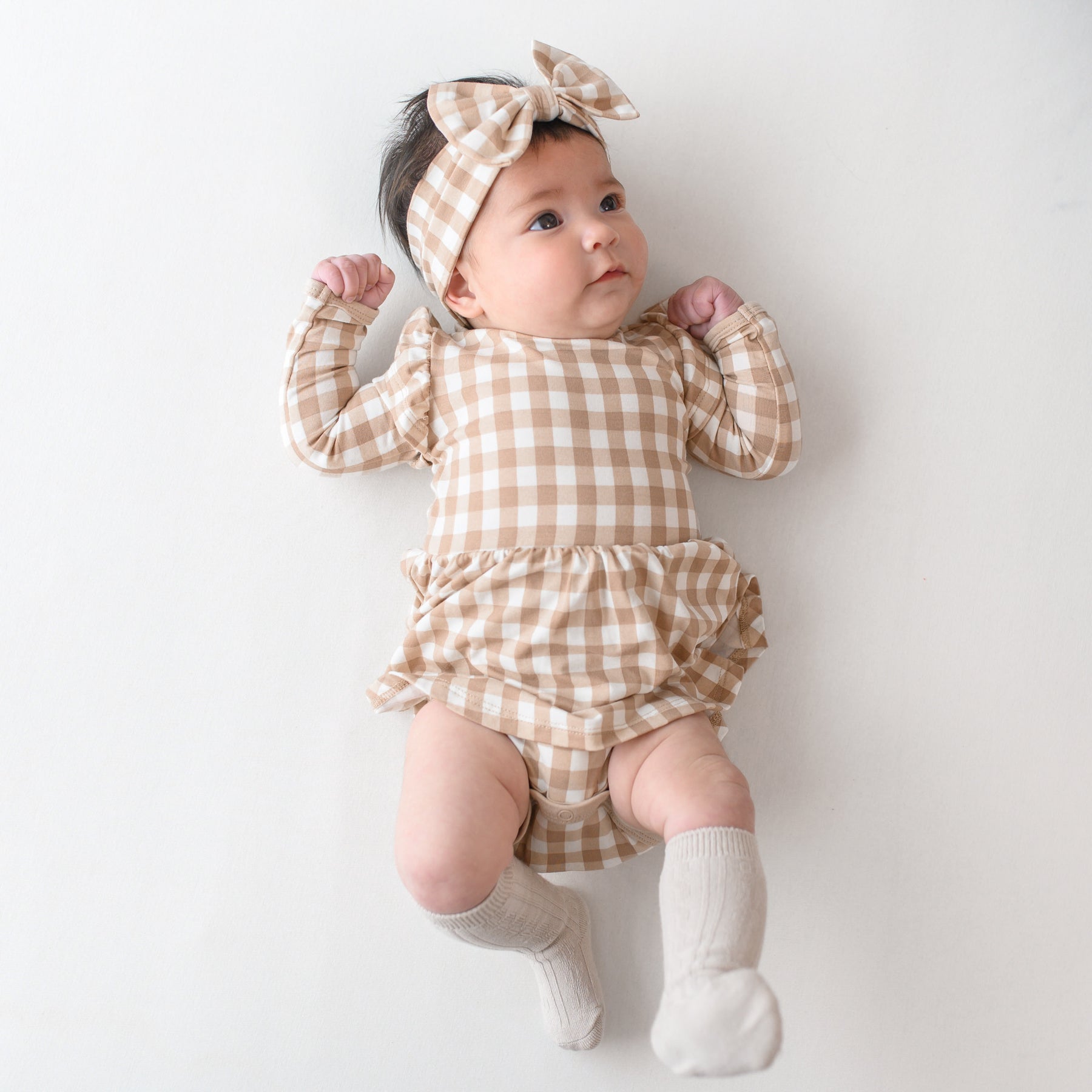 Infant girl laying on a light colored neutral backdrop looking off to the side wearing the Long Sleeve Twirl Bodysuit Dress in Gingham Bisque and matching bow headband with Oat Knee high socks
