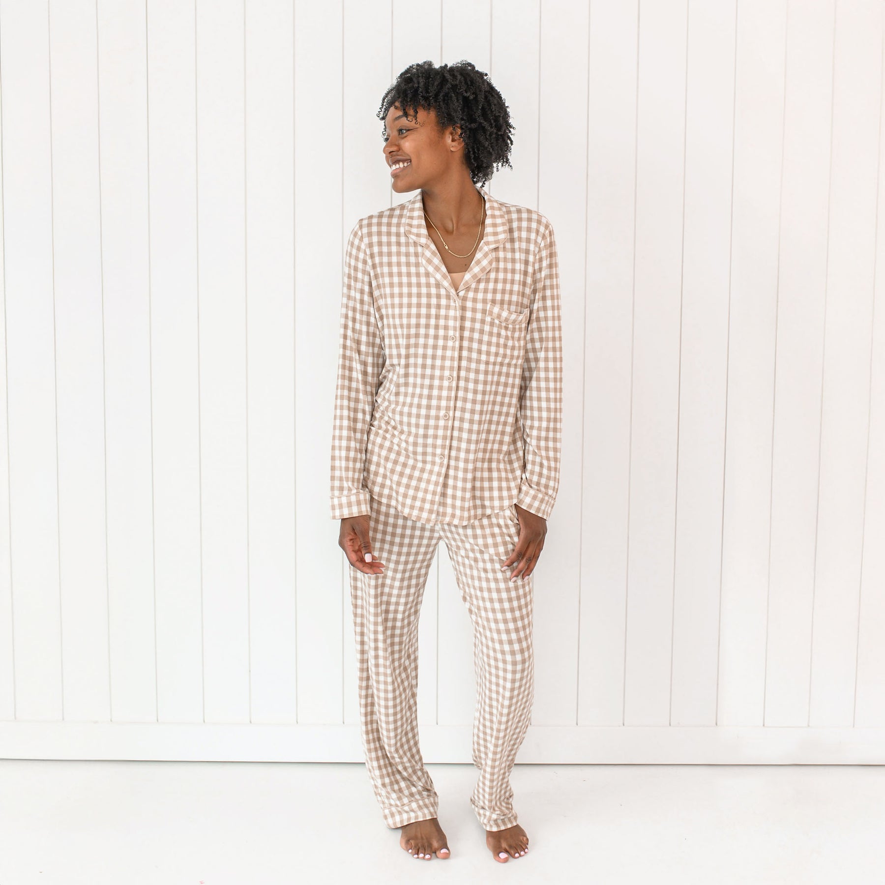 Female model wearing the Long-Sleeved Women's Pajama Set in Gingham Bisque standing in front of a white paneled wall with one hand in the pajama pant pocket