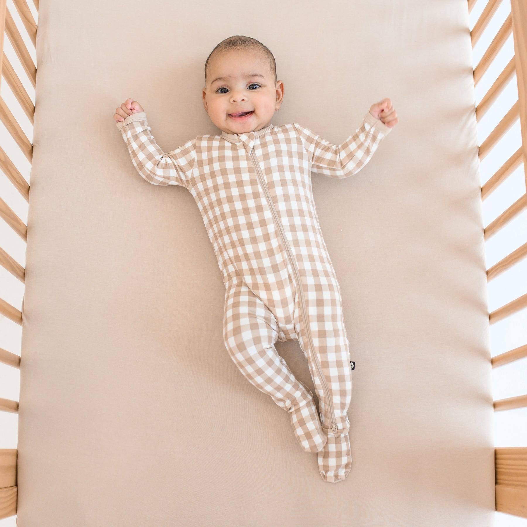 Infant laying in a crib on a Bisque crib sheet wearing the Zippered Footie in Gingham Bisque