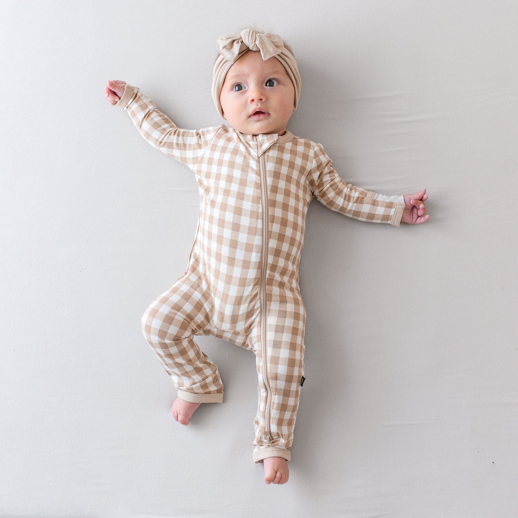 Infant laying down wearing the Zippered Romper in Gingham Bisque and Knotted Bow headband in Bisque