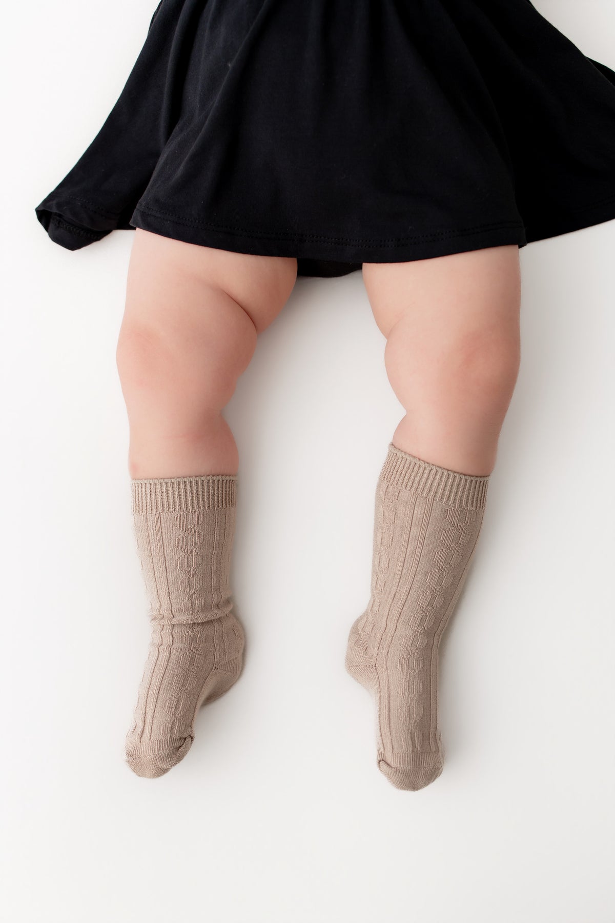 A close-up of a child's legs wearing ribbed knee-high socks in a taupe beige color, paired with a black skirt, against a plain white background.