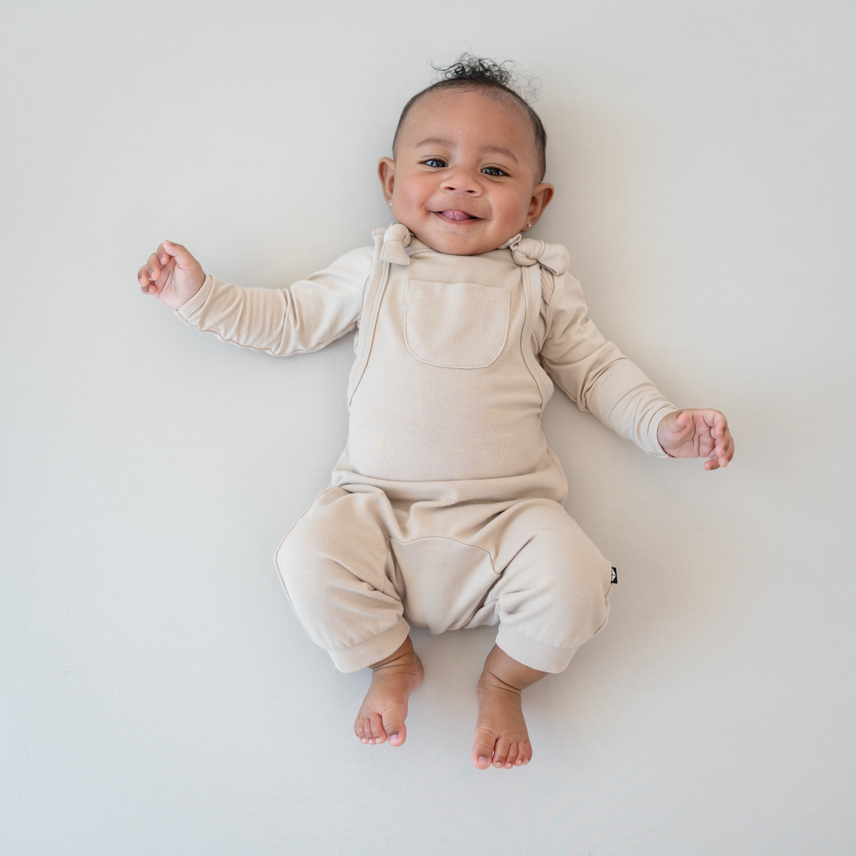 Smiling infant laying down with tongue sticking out wearing the Bamboo Jersey Overall in Bisque with matching long sleeve bodysuit on underneath