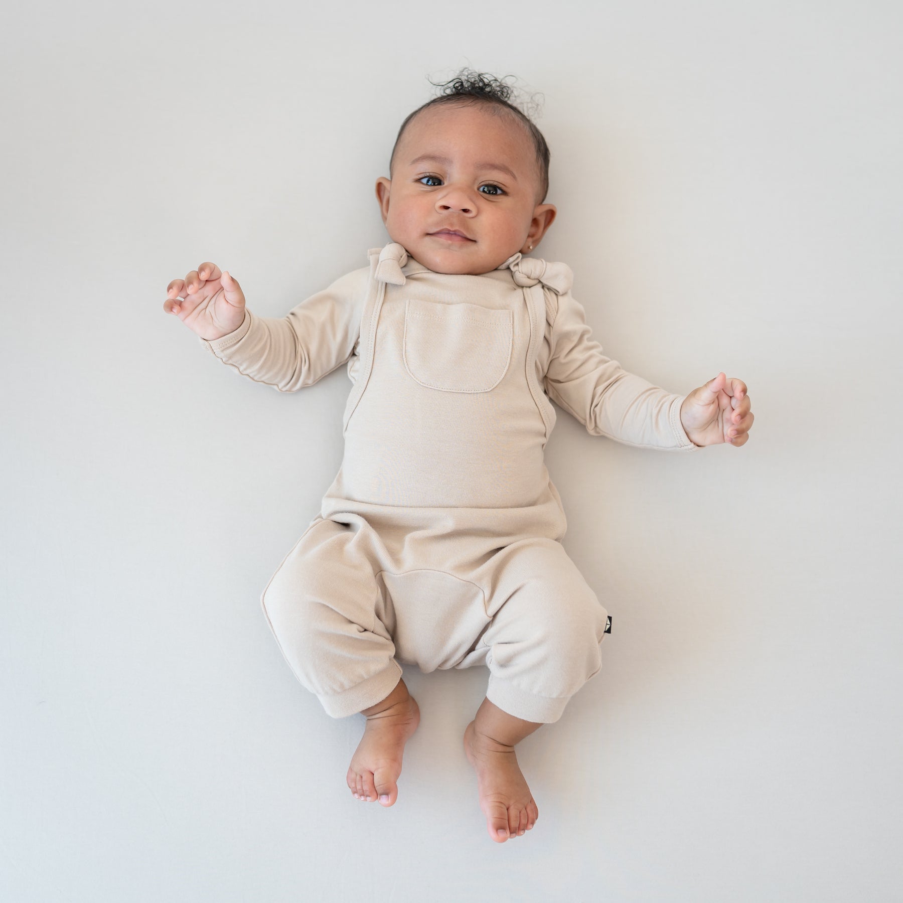 Infant laying down wearing the Bamboo Jersey Overall in Bisque with matching long sleeve bodysuit on underneath