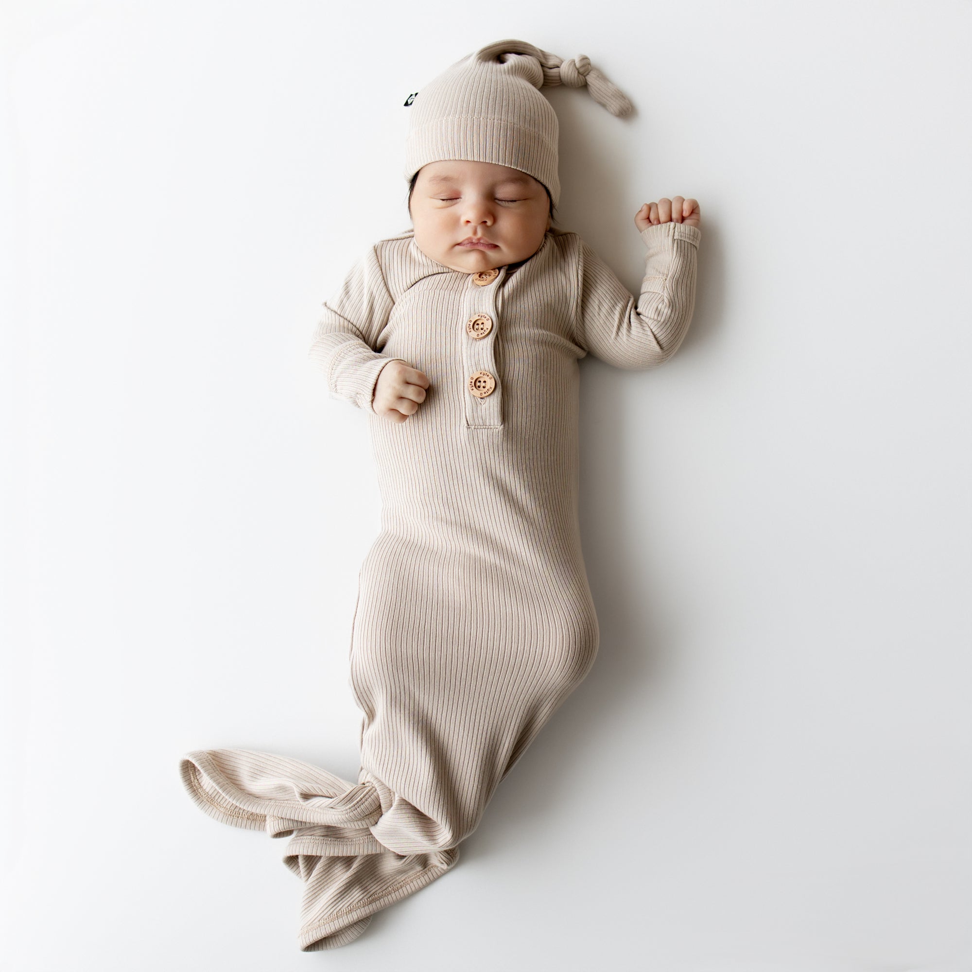 Sleeping infant wearing the Ribbed Knotted Gown with Hat Set in Bisque laying on a white backdrop
