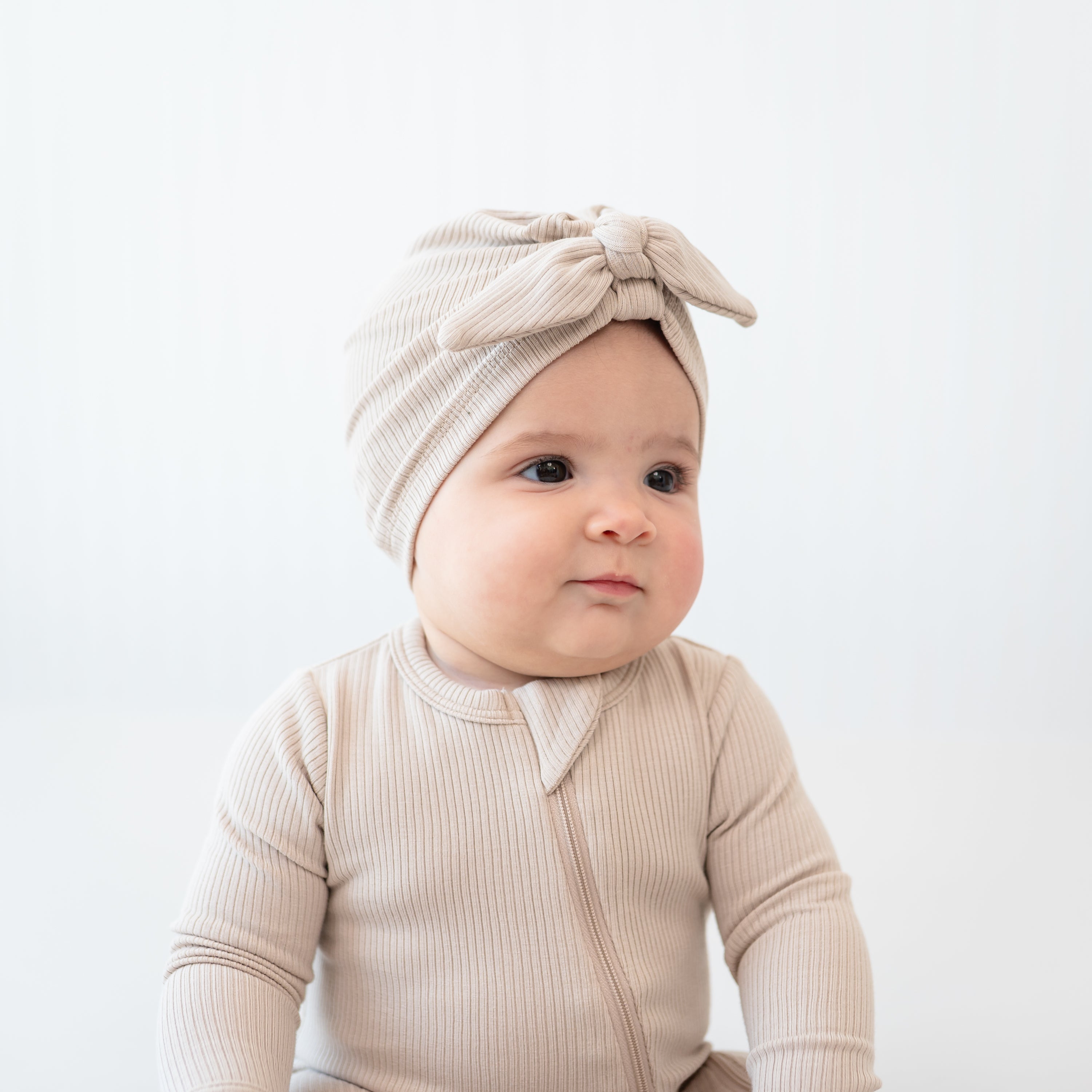 Close up of an infant wearing the Ribbed Headwrap in Bisque with matching zippered romper in front of  a white back drop