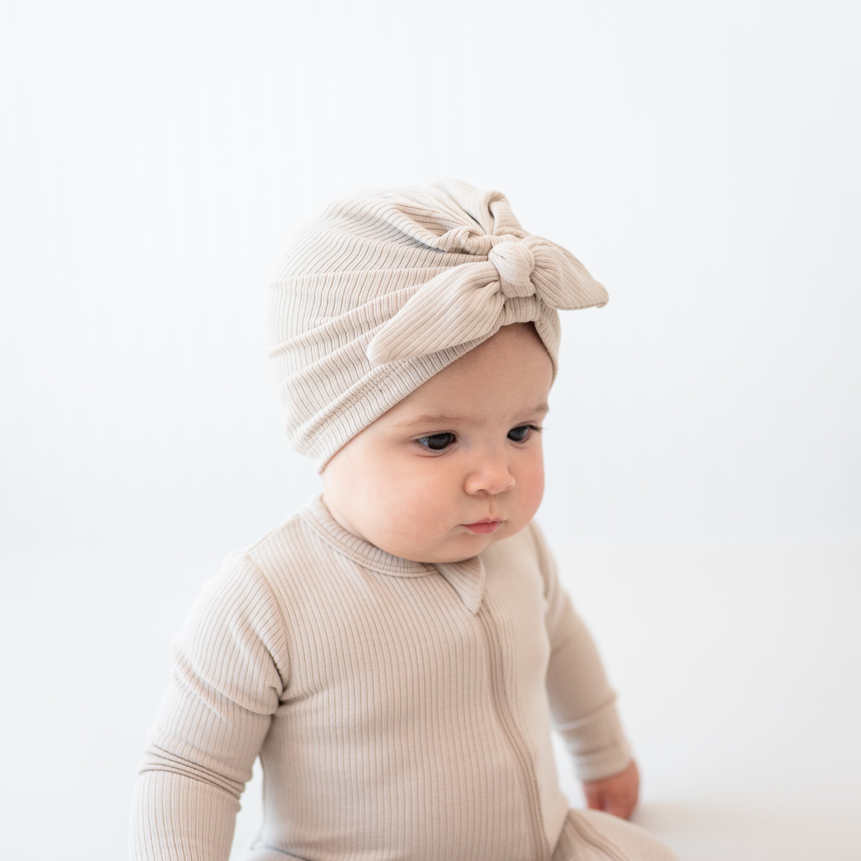 Top side view of the Ribbed Headwrap in Bisque on a sitting infant model wearing a matching ribbed zipper footie in front of a white backdrop