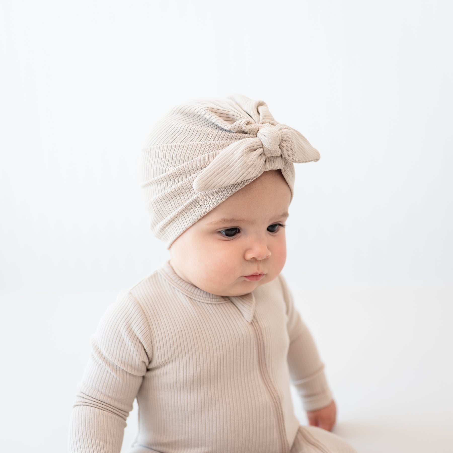Top side view of the Ribbed Headwrap in Bisque on a sitting infant model wearing a matching ribbed zipper footie in front of a white backdrop