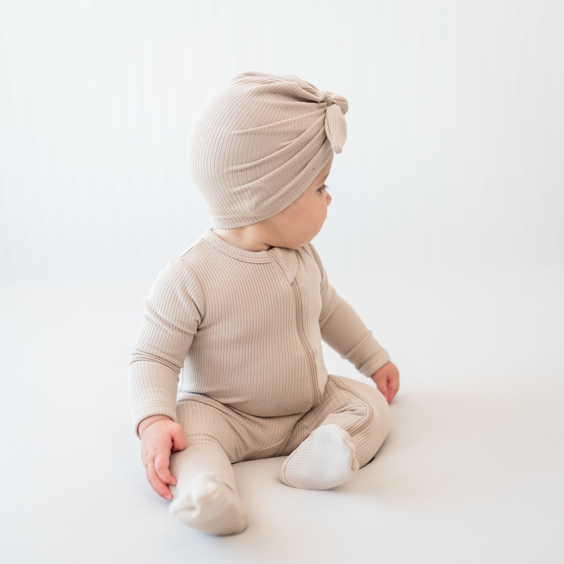 Side view of the Ribbed Headwrap in Bisque shown on an infant model wearing the Bisque zippered romper in front of a light backdrop
