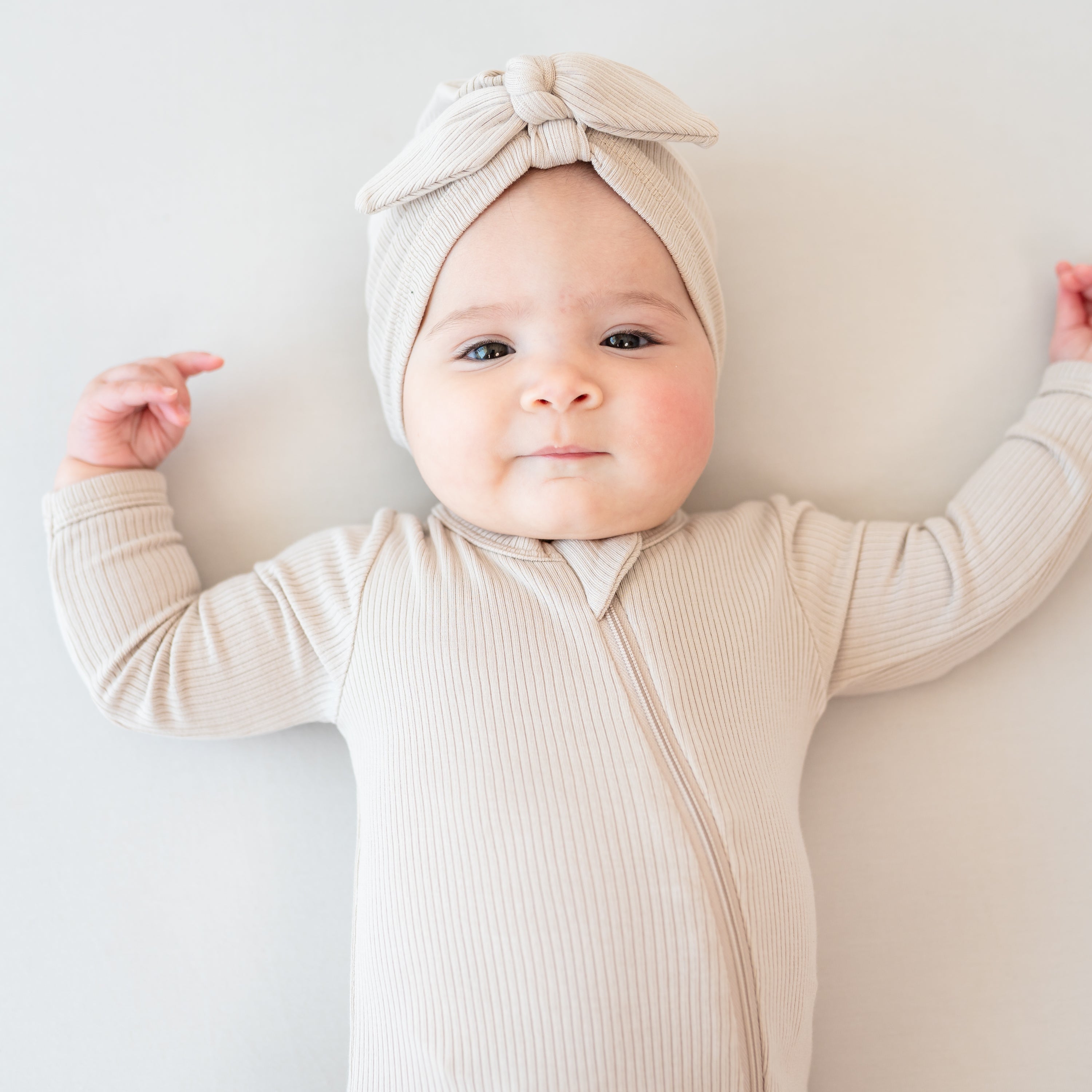 Infant lying down on a Bisque blanket wearing the Ribbed Headwrap in Bisque with matching ribbed zipper romper