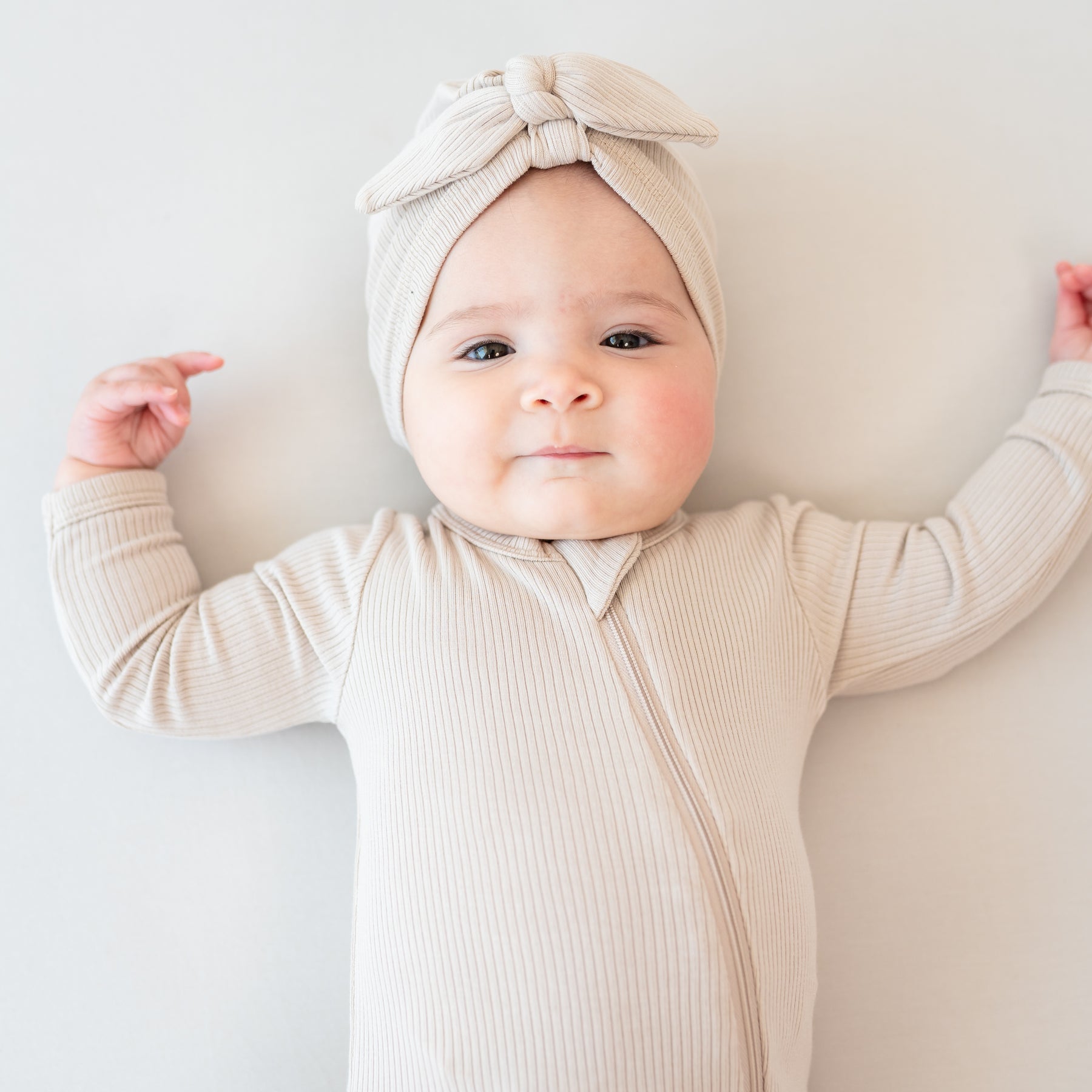 Infant lying down on a Bisque blanket wearing the Ribbed Headwrap in Bisque with matching ribbed zipper romper