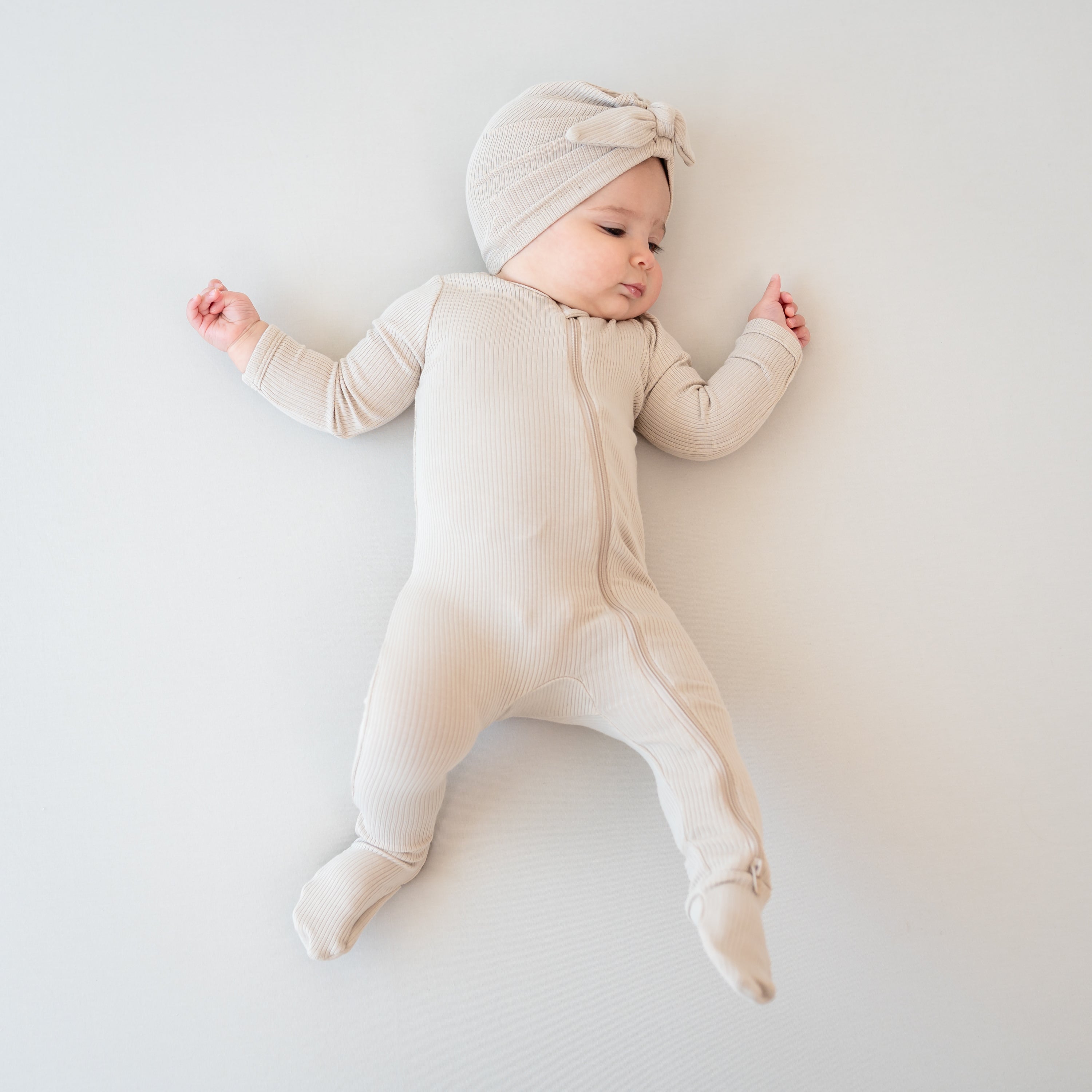 Infant girl laying down looking to the side wearing the Ribbed Zipper Footie in Bisque paired with a matching ribbed headwrap