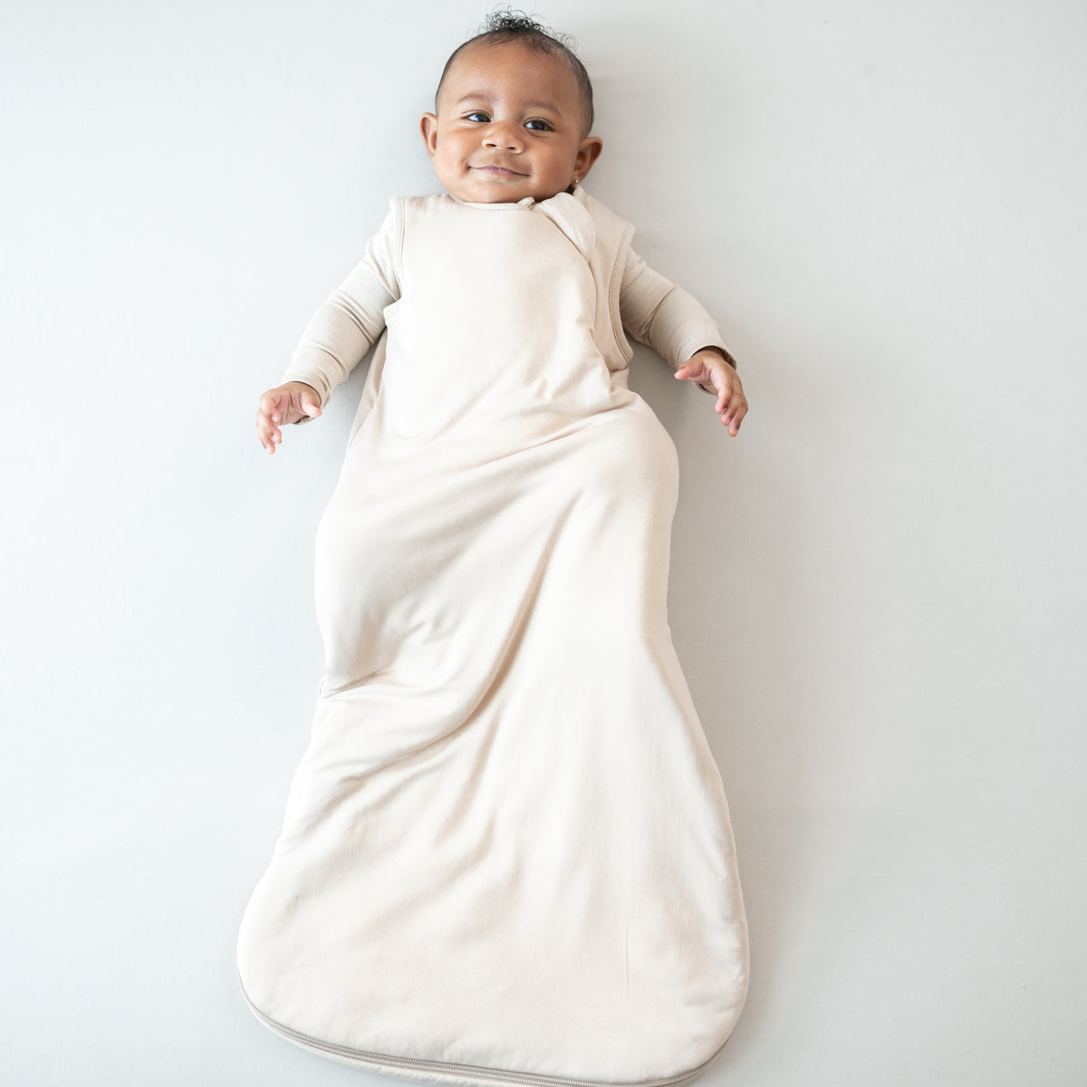 Infant laying down wearing the Sleep Bag in Bisque 1.0 with matching romper on underneath