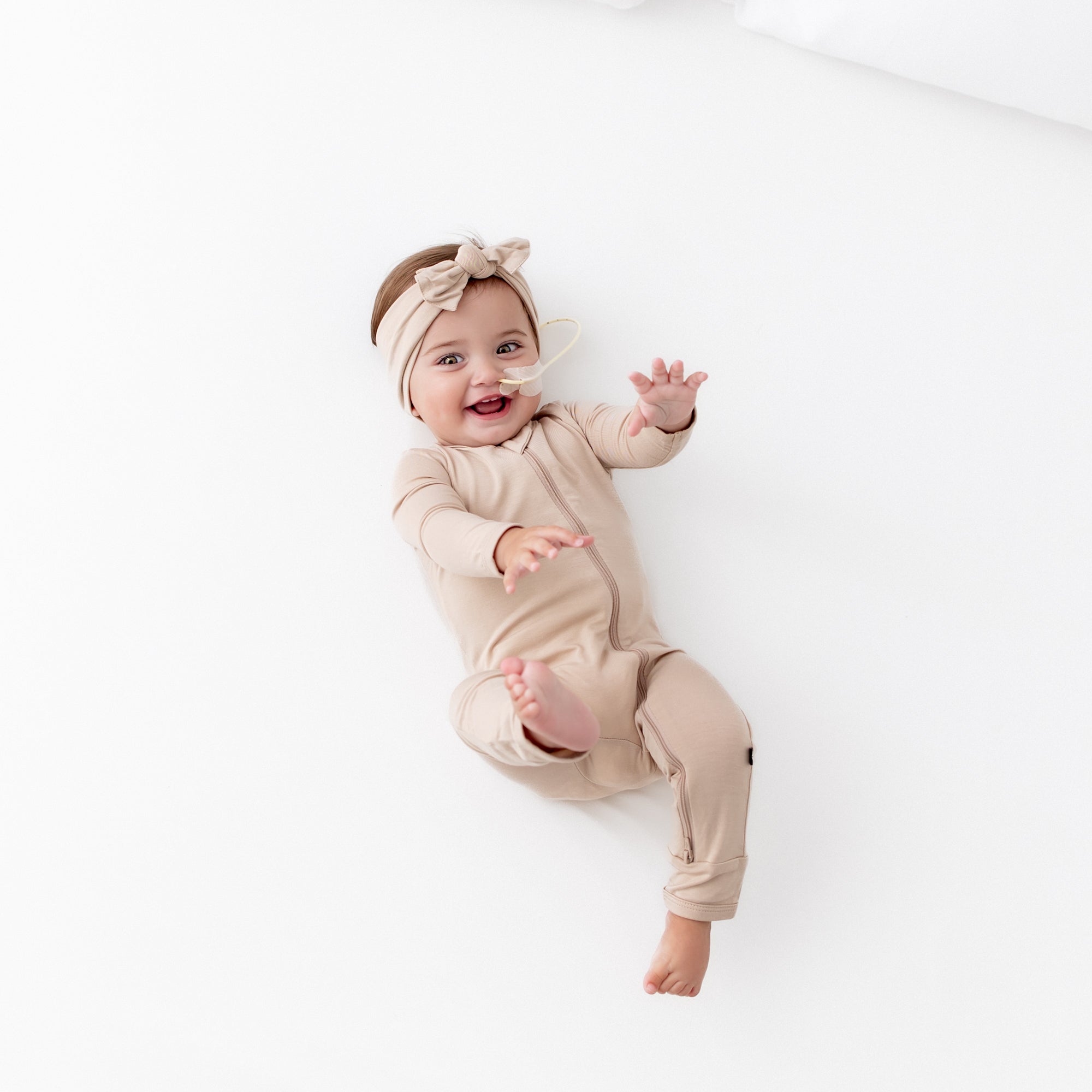 Smiling infant wearing the Zippered Romper in Bisque with matching knotted bow headband