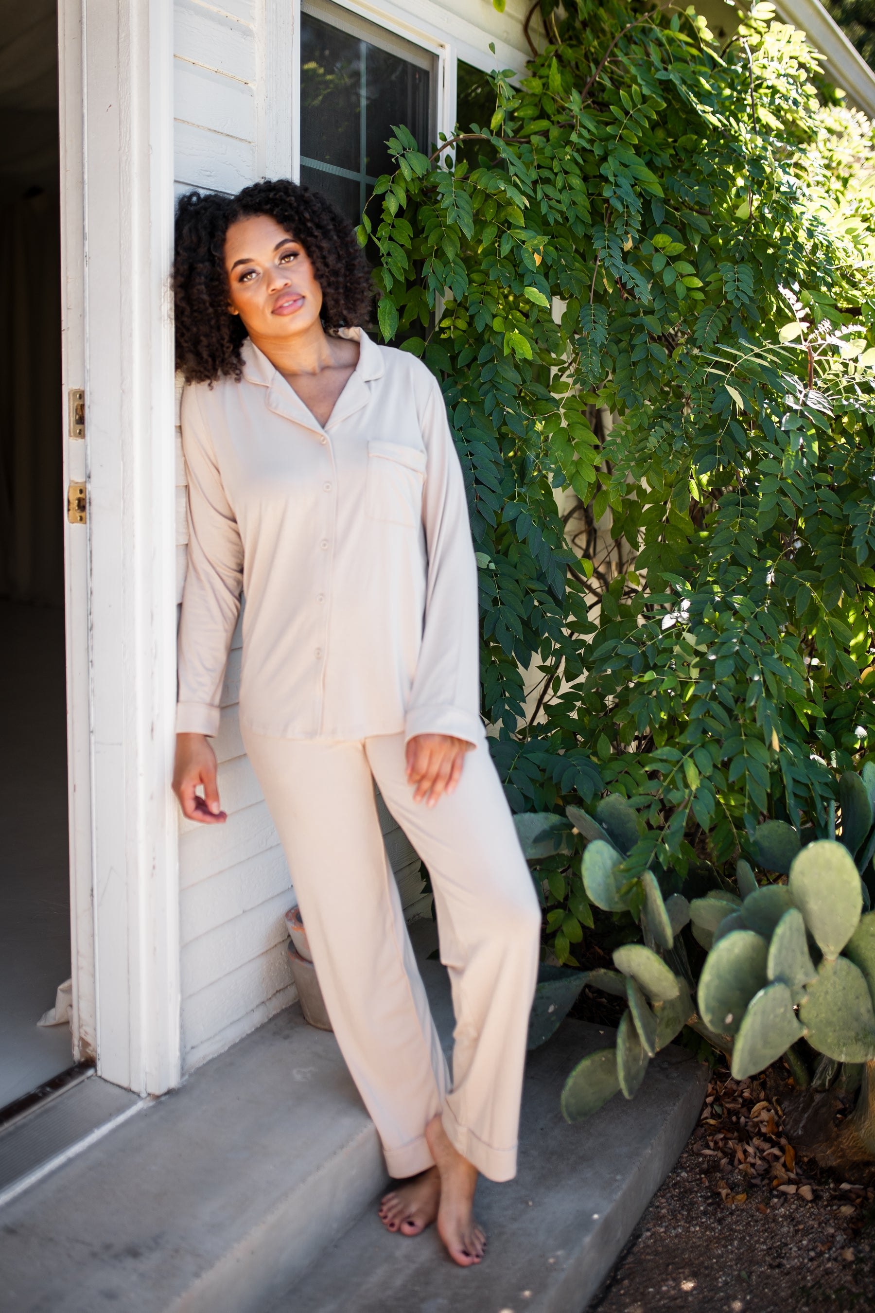 Female model leaning against a house wearing the Long-Sleeved Women's Pajama Set in Bisque with Bisque Trim