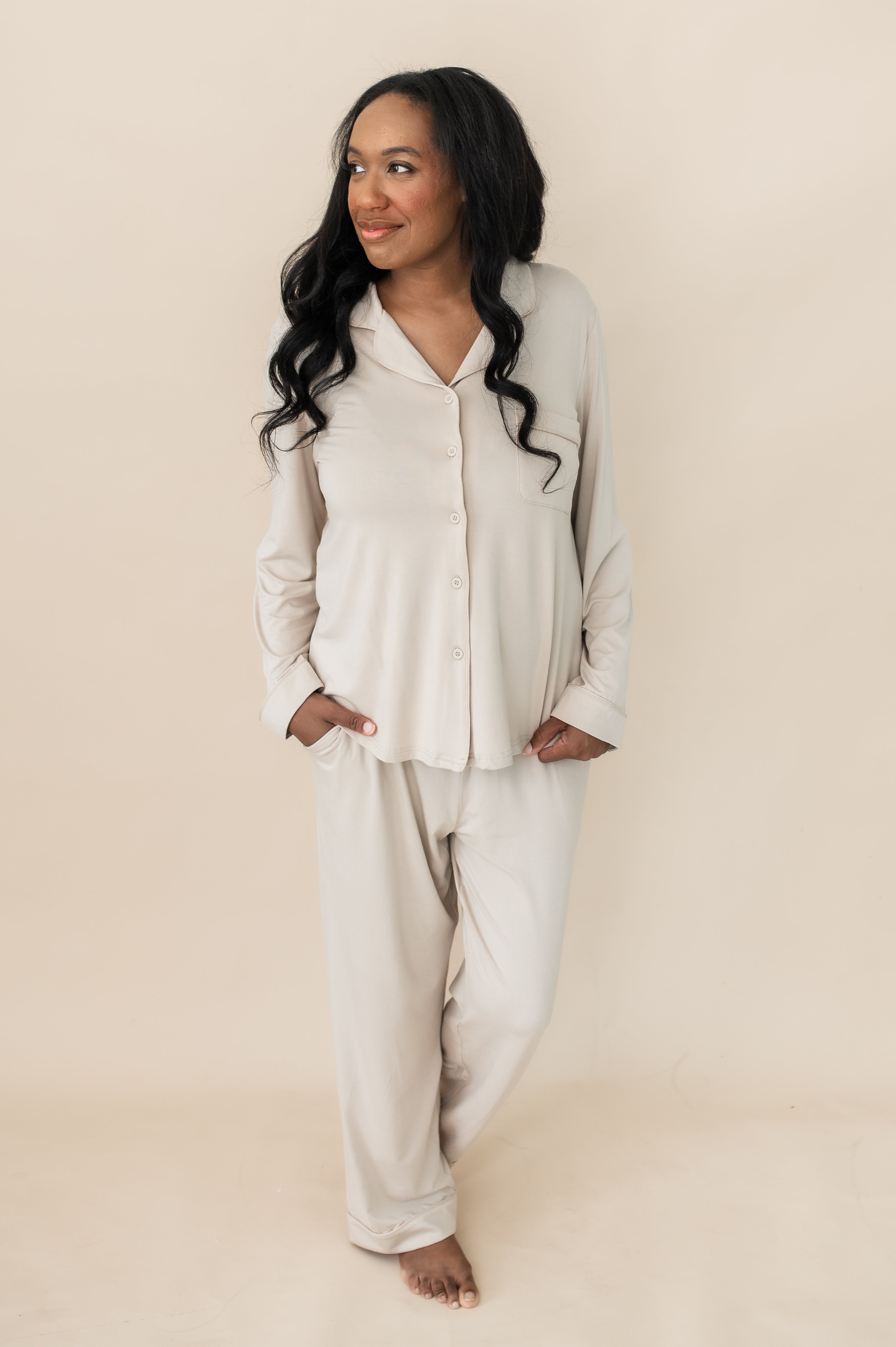 Female modeling the Long-Sleeved Women's Pajama Set in Bisque with Bisque Trim with one hand in the pants pocket and the other holding the bottom hem of the shirt