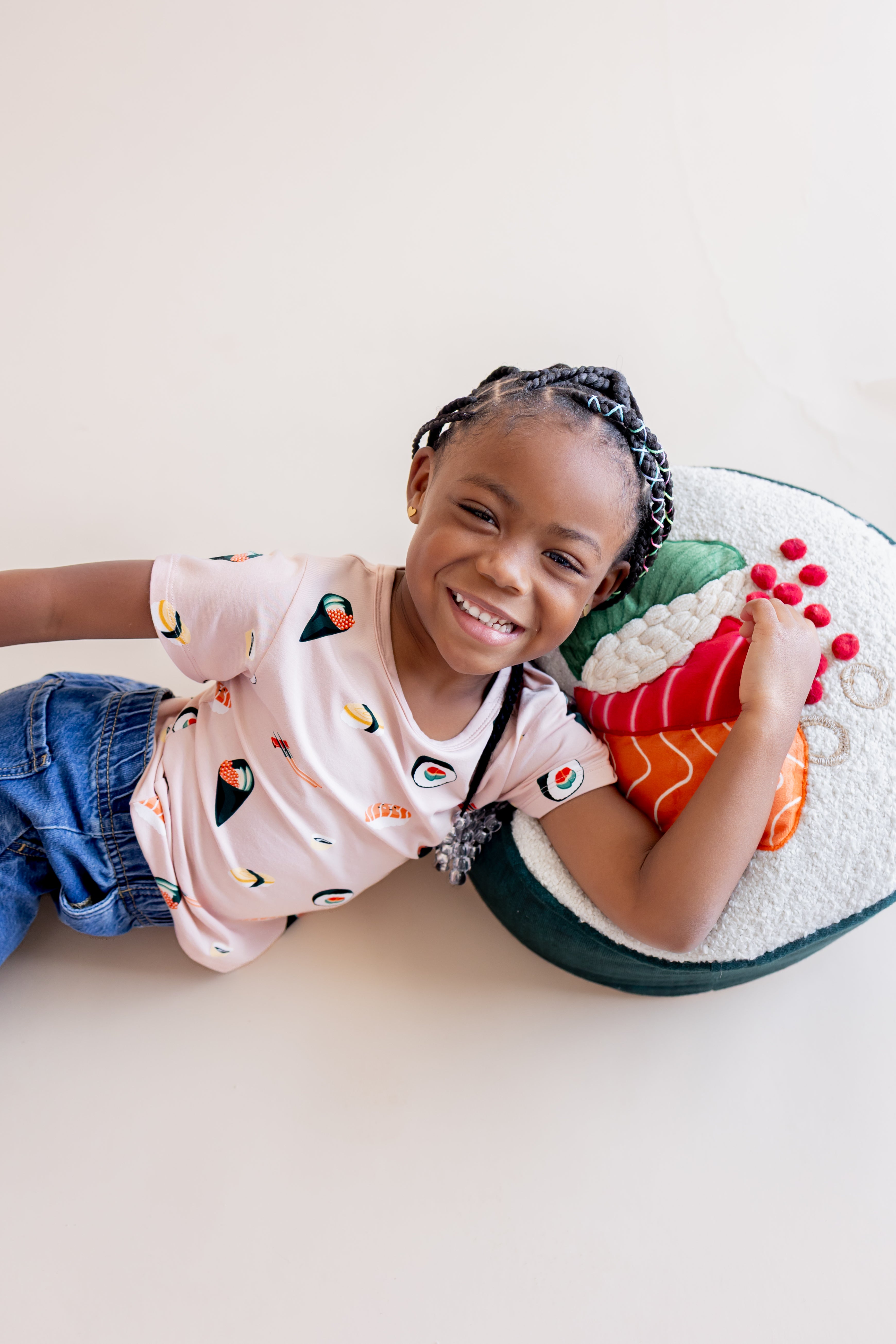 Child smiling, laying on their side in blush sushi tee, jeans, and a play sushi pillow