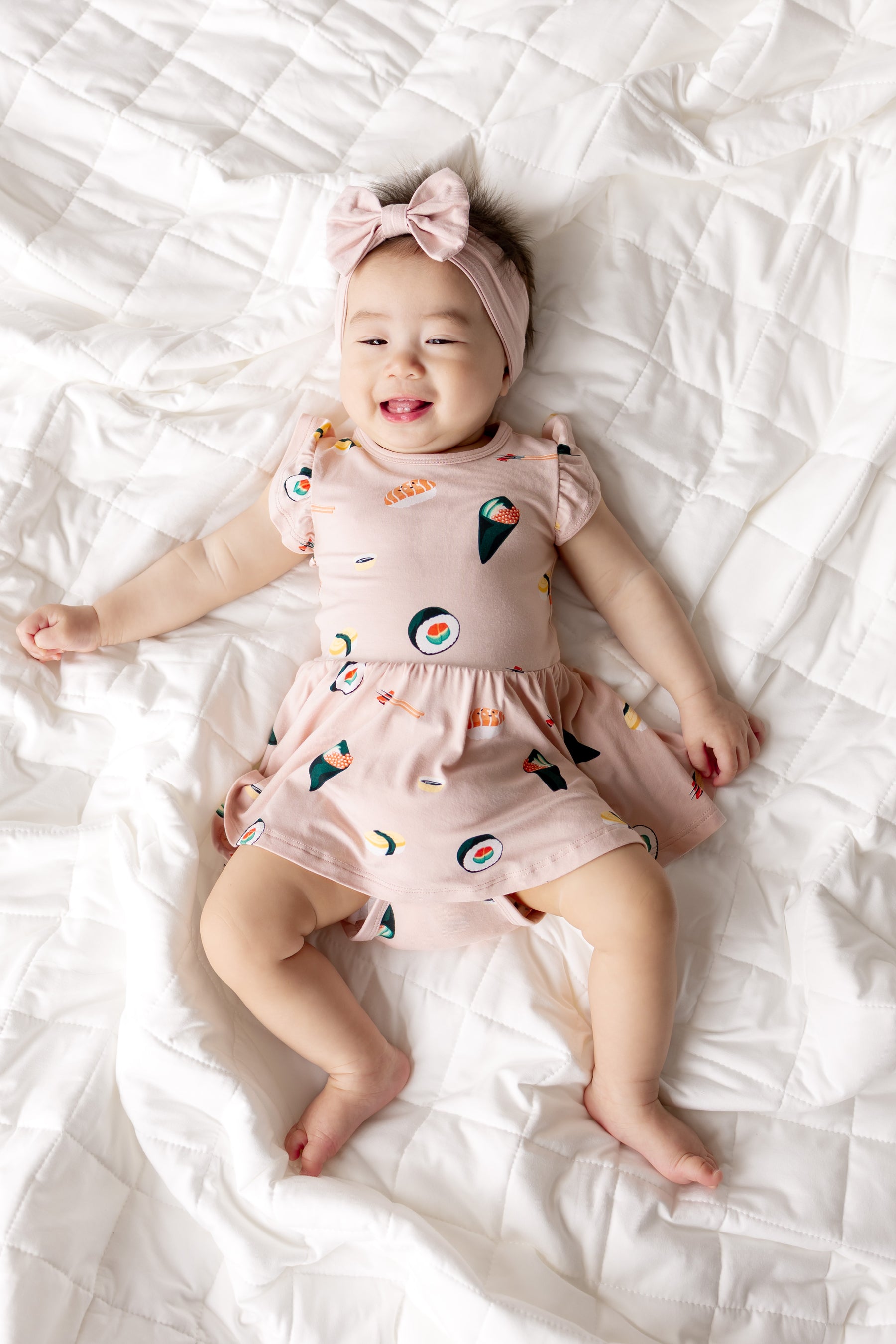 Baby wearing Pink Twirl Bodysuit Dress with Sushi print on it and pink headband on the head lying on white sheets