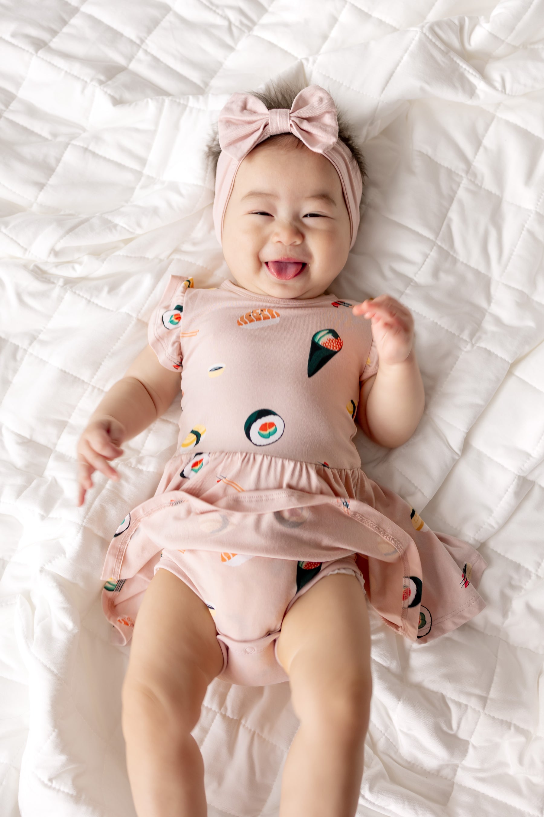 Baby smiling in Pink Twirl Bodysuit Dress with Sushi print and pink headband on the head lying on white sheets